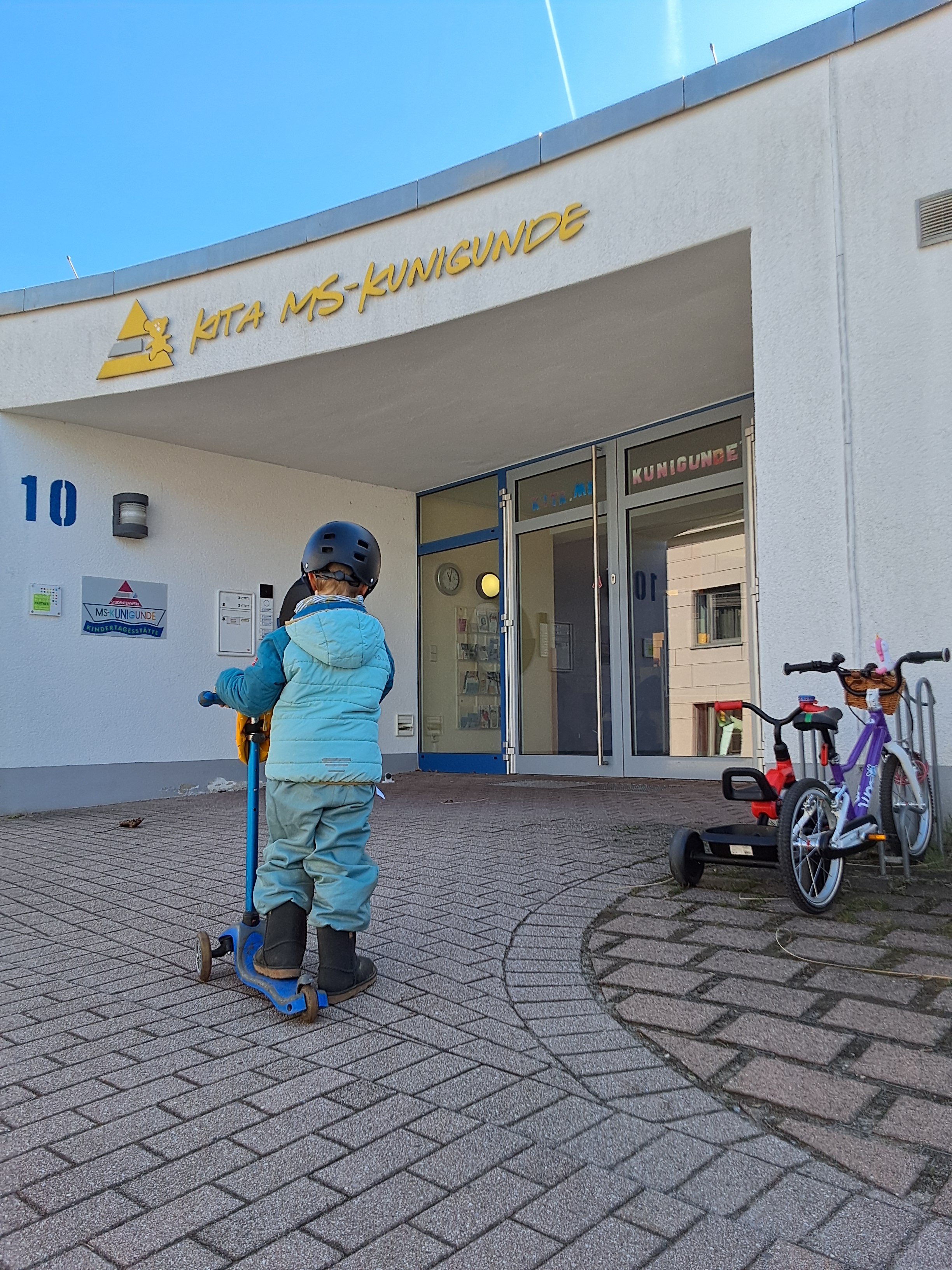 Ein Kind steht auf einem Roller. Im Hintergrund ist ein Eingang zum Gebäude mit dem Schriftzug "Kita MS-Kunigunde" zu erkennen.