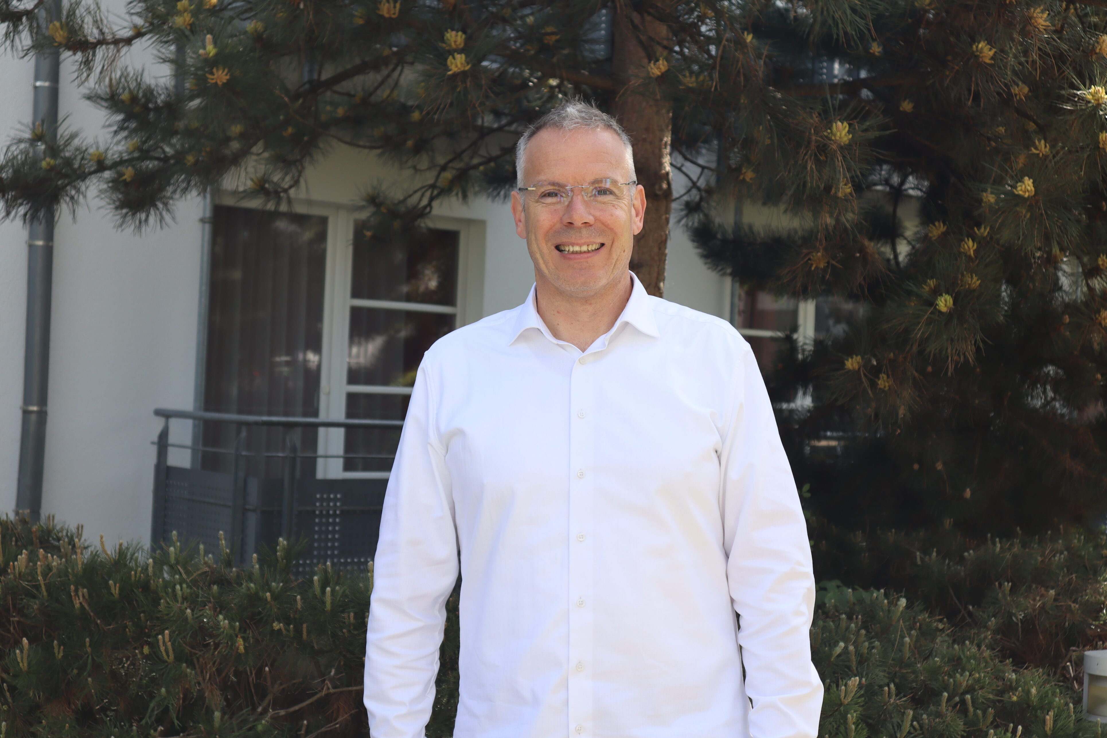Eine Person steht vor einem Baum. Sie hat kurze Haare, trägt eine Brille und ein weißes Hemd.