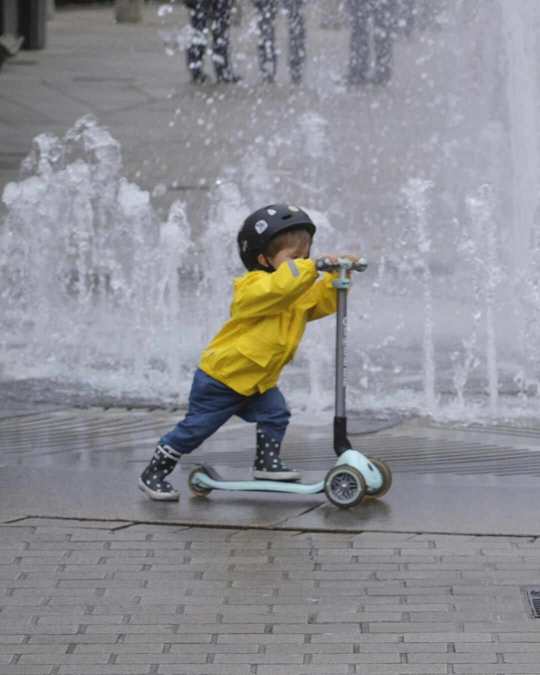 A child with a yellow rain jacket and helmet is riding a scooter.