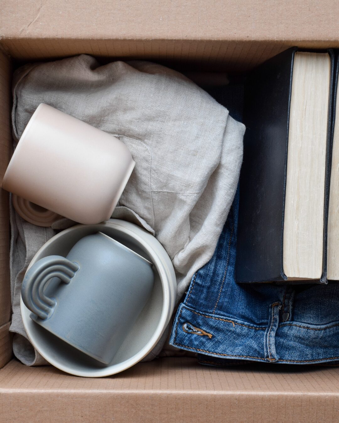 Ein Karton, in dem Bücher, Kleidung und zwei Tassen liegen.
