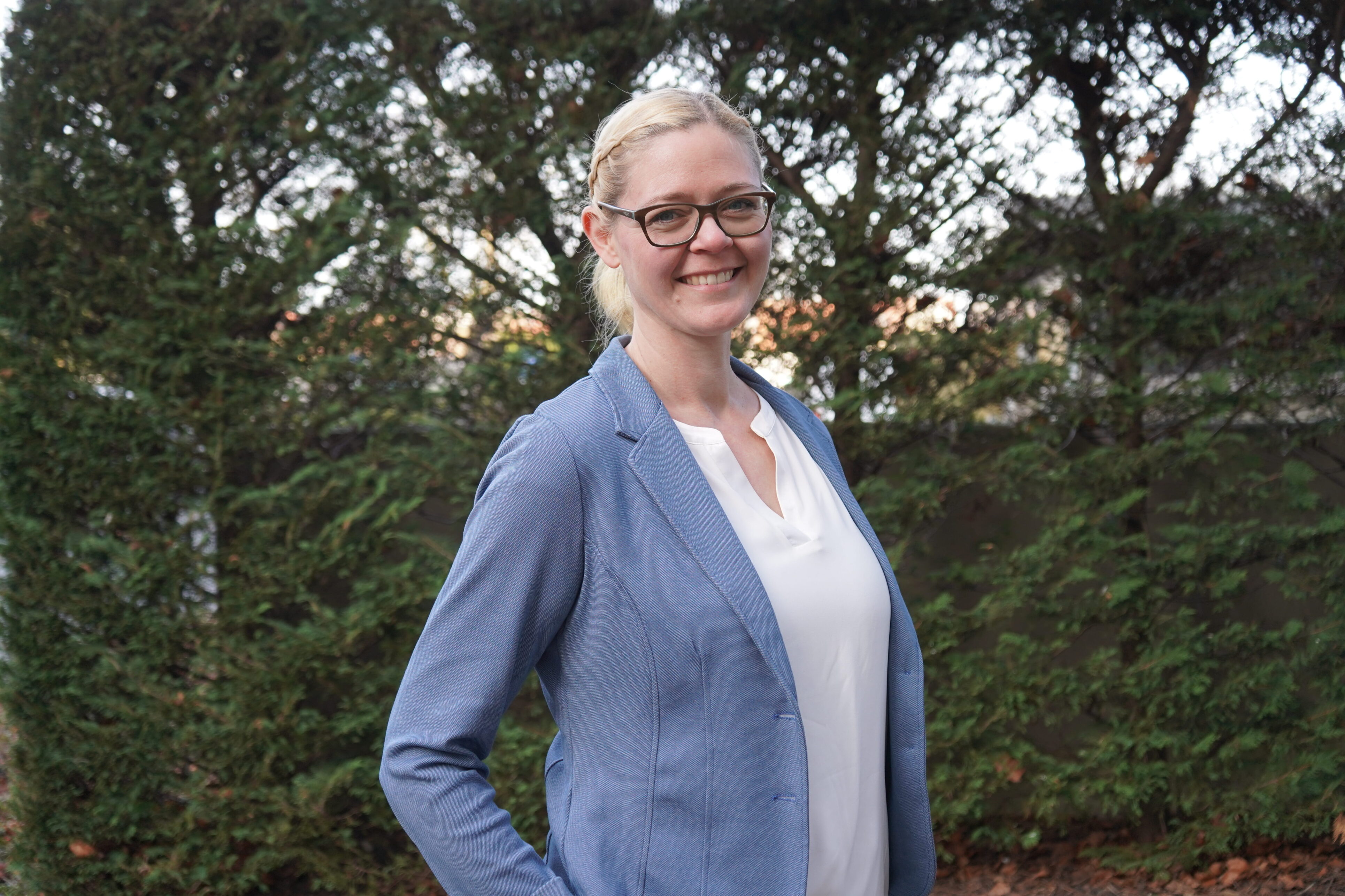 Person mit blonden mittellangen Haaren, die in einem Zopf gebunden sind, einer Brille und einem blauen Blazer. Sie steht vor einem grünen Hintergrund.