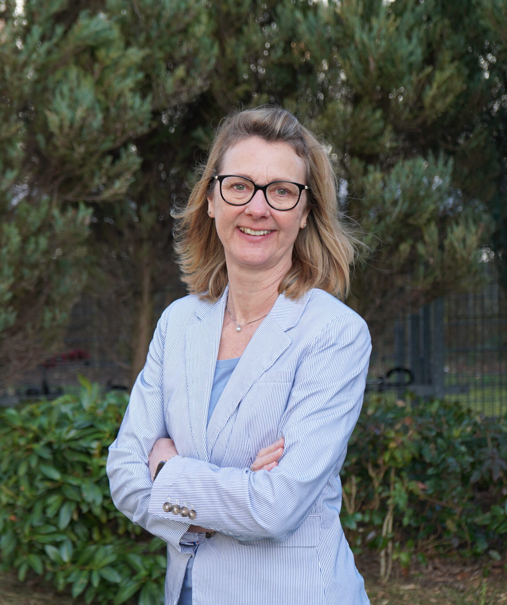 Eine Person mit schulterlangen Haaren und Brille hat die Arme vor der Brust verschränkt und lächelt in die Kamera. Sie trägt einen blau-weiß gestreiften Blazer und steht vor einer grünen Hecke.