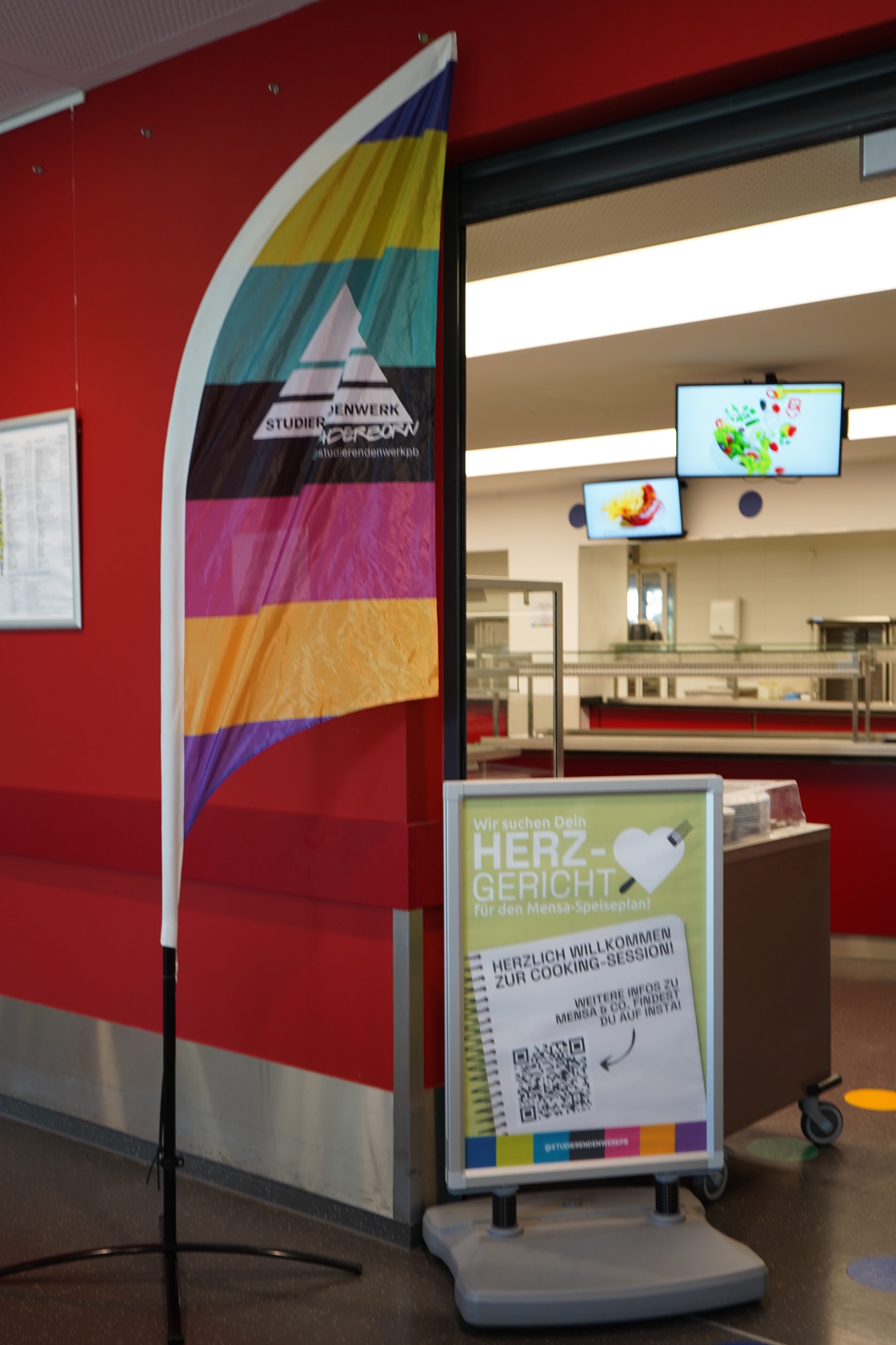 In the entrance area of a canteen, there is a beach flag and an A1 stand bearing the words "Welcome to the Cooking Session!"