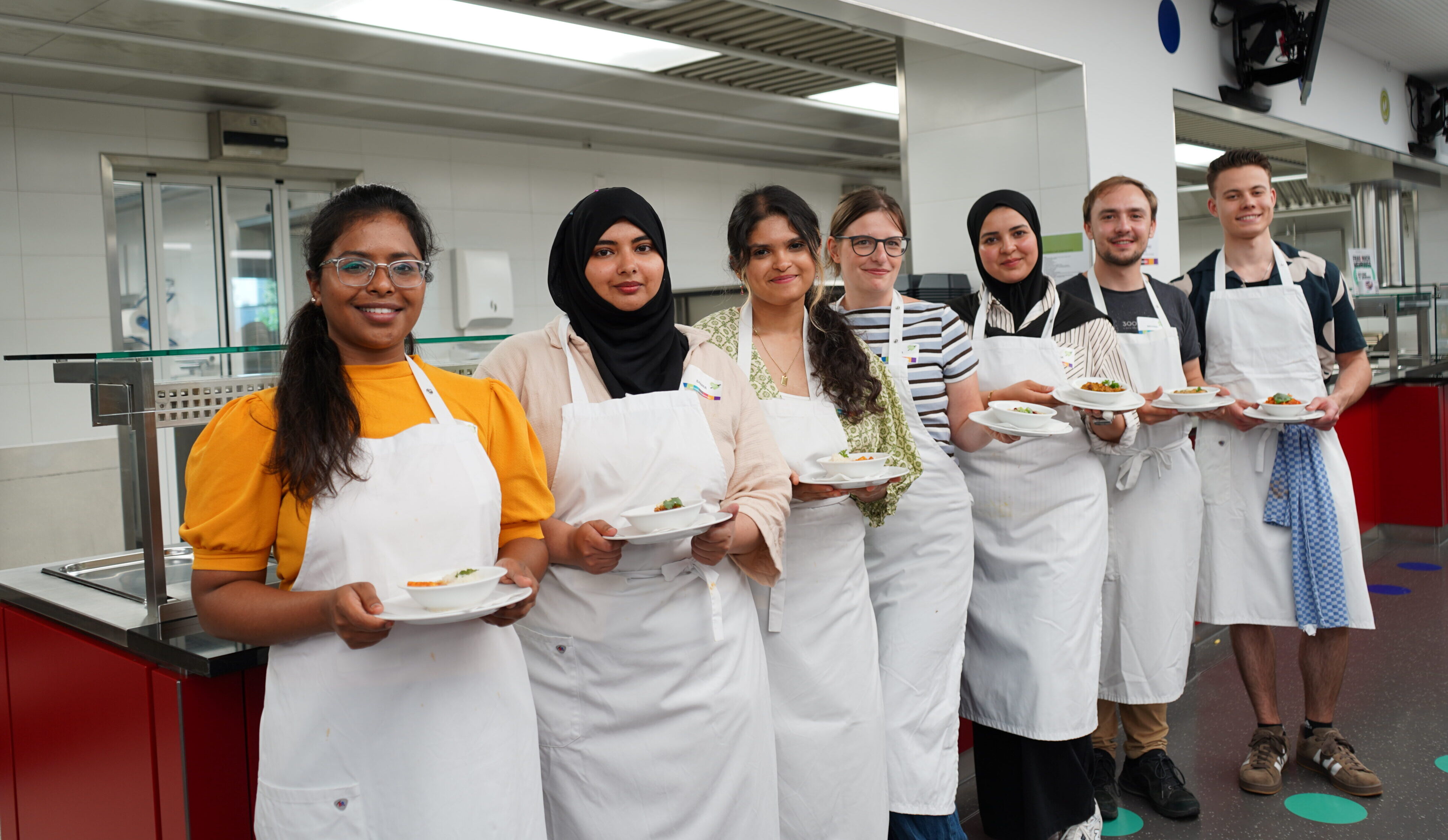 Seven people are standing in a row in a kitchen, looking at the camera. They are wearing aprons and each has a plate in their hand.