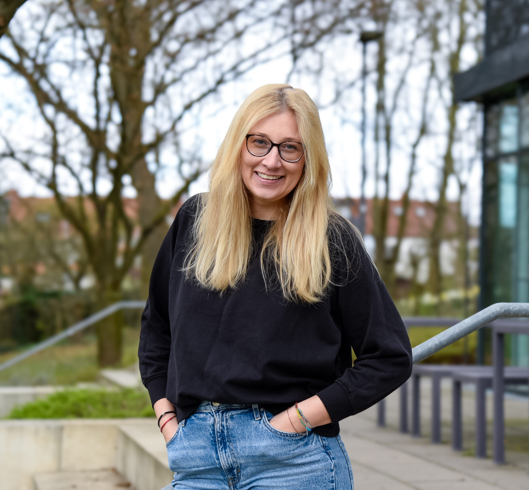 Eine Person mit langen blonden Haaren, einer Brille und einem schwarzen Pullover steht vor einem gläsernen Gebäude, umringt von Bäumen.