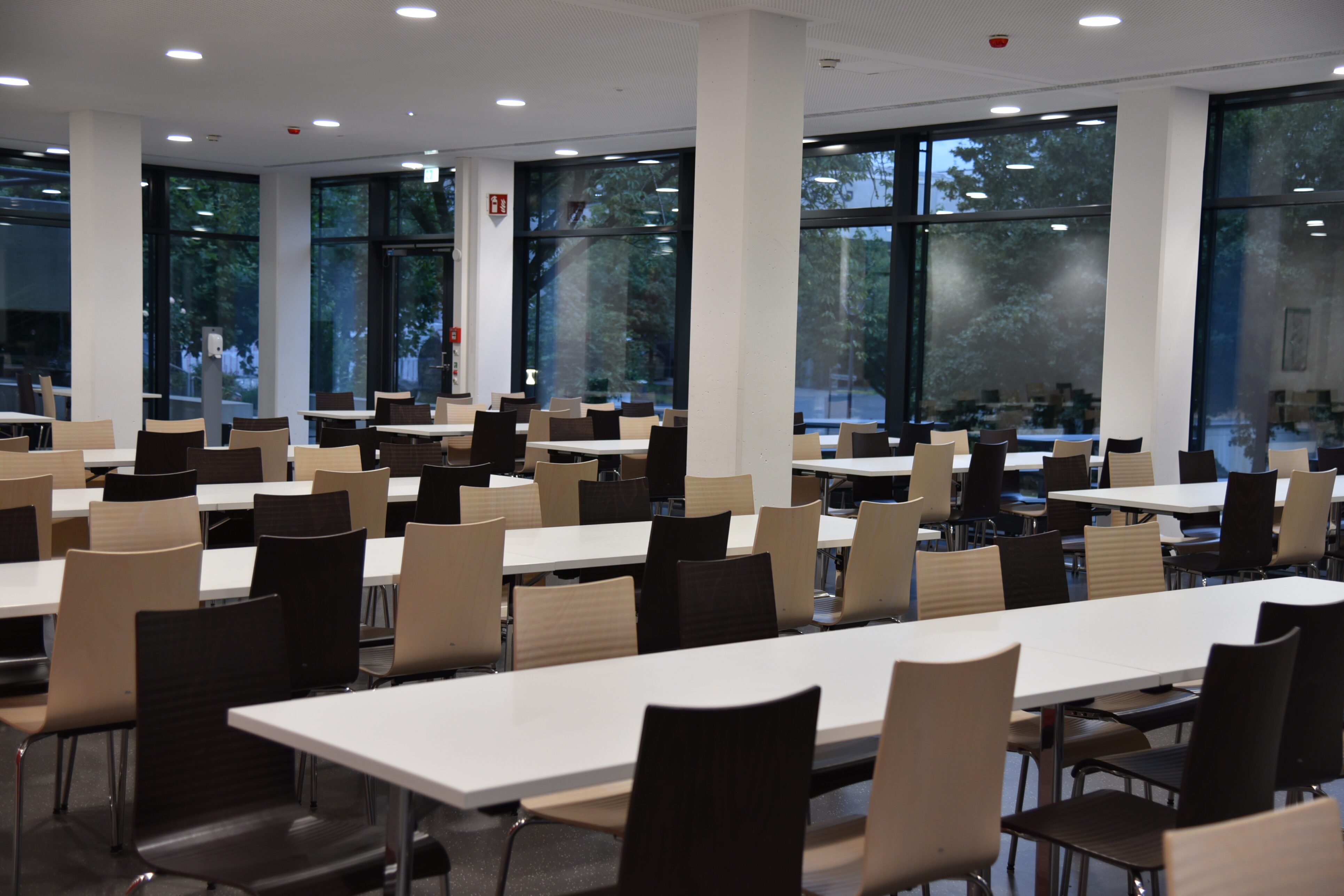 Offener Sitzbereich mit Tischen und Stühlen umgeben von einer großzügigen Glasfassade in einer Mensa