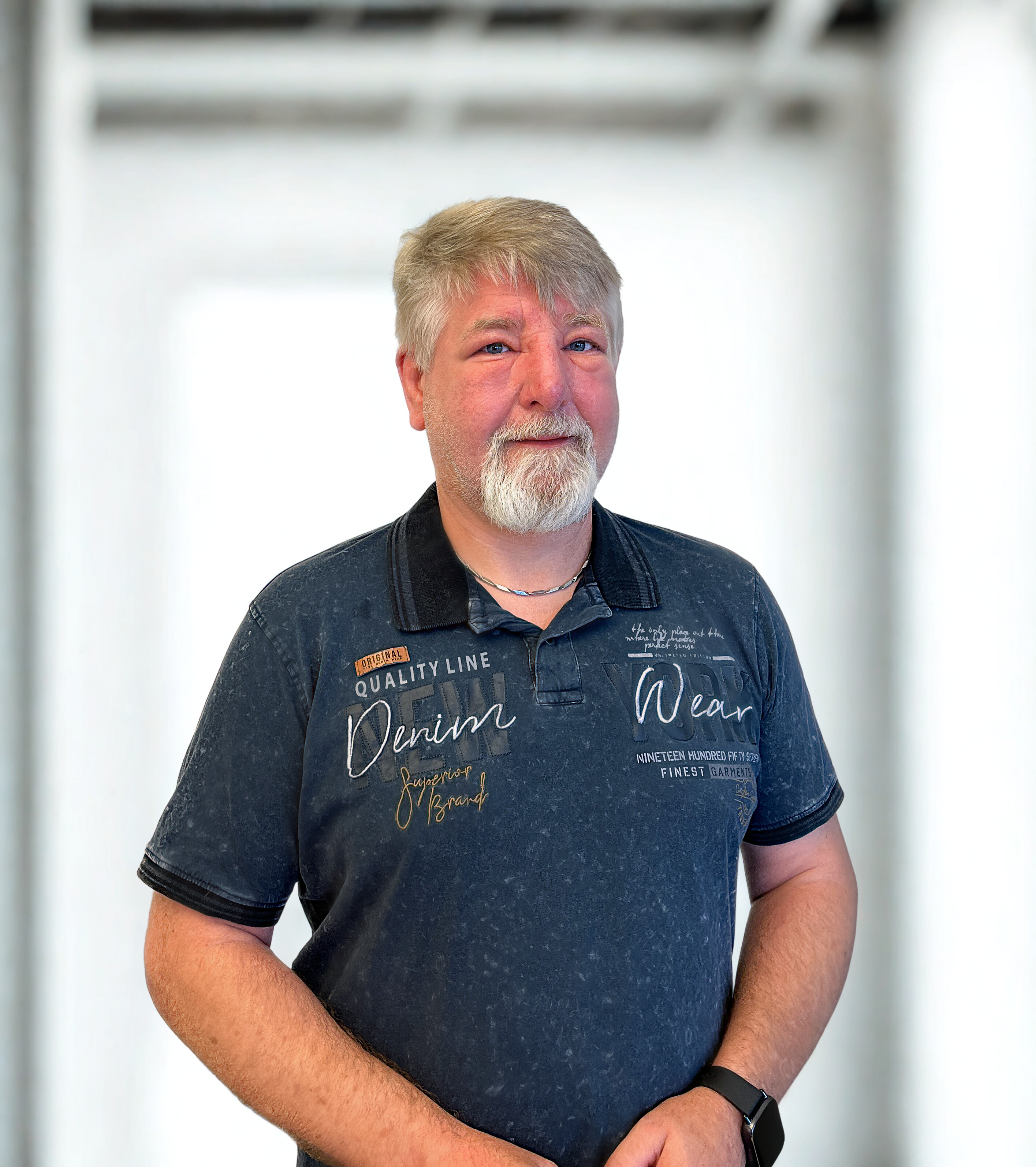 Eine Person steht in einem hellen Raum. Sie hat kurze Haare, einen Bart und trägt ein Poloshirt.