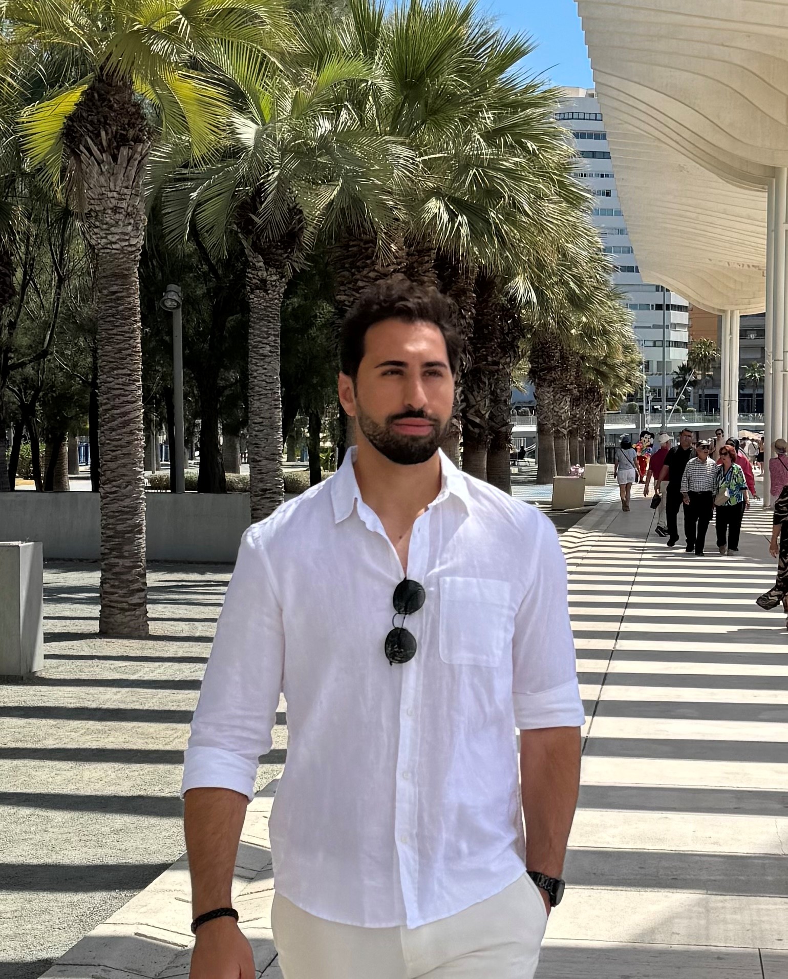 Im Mittelpunkt steht eine Person, die auf die Kamera zuläuft. Die Sonne scheint und im Hintergrund sind Palmen. Die Person hat einen Bart und trägt ein lockeres weißes Hemd.