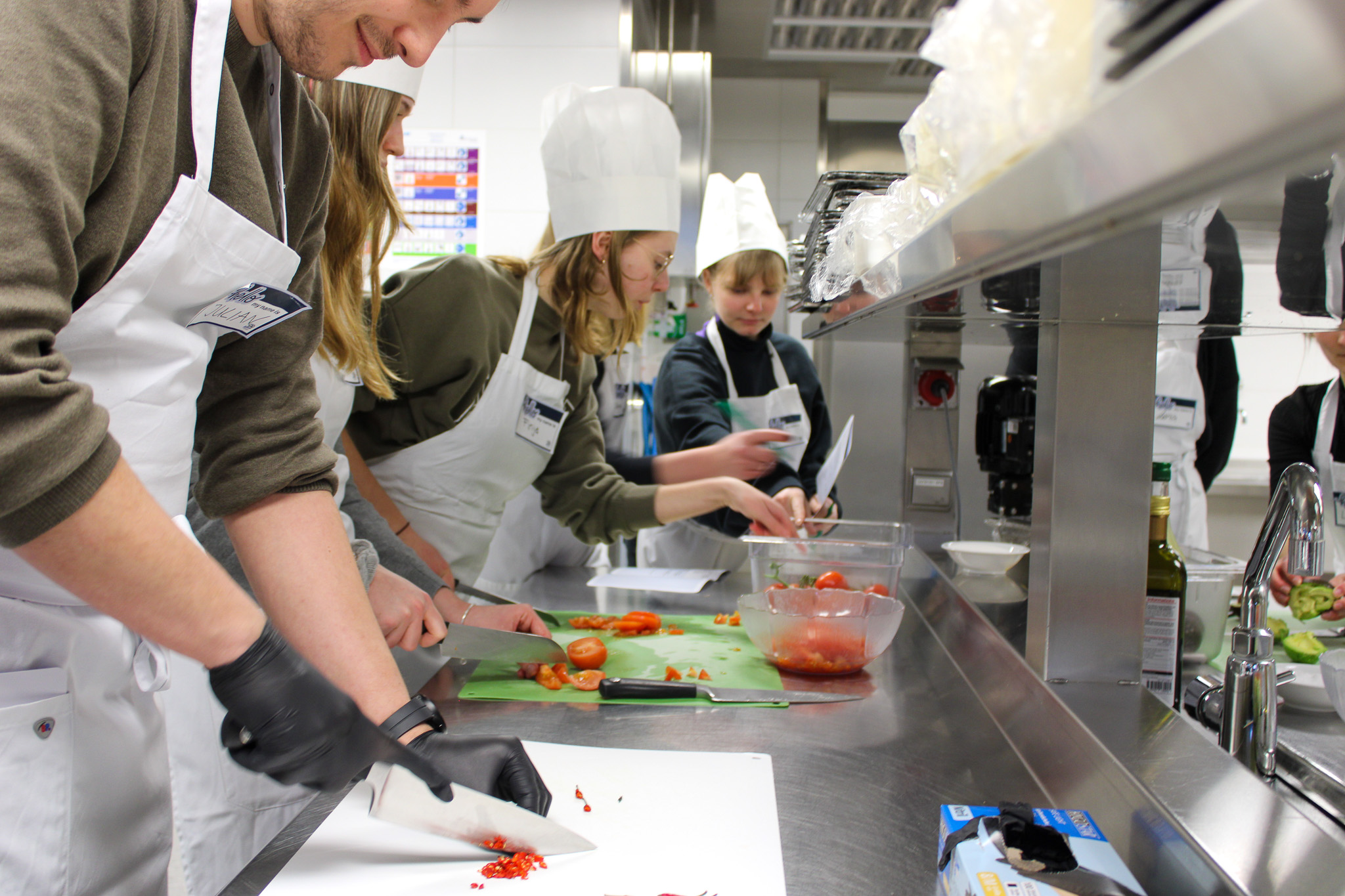 Drei Personen mit Schürze und Hut tragen Handschuhe und stehen vor einer silberfarbenen Arbeitsfläche und schneiden Gemüse.