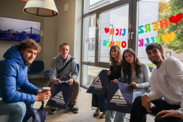 Personen sitzen im Halbkreis nebeneinander und schauen in die Kamera. Sie halten blaue Jute-Beutel in der Hand und hinter ihnen hängt eine Girlande mit der Aufschrift "Herzlich Willkommen"