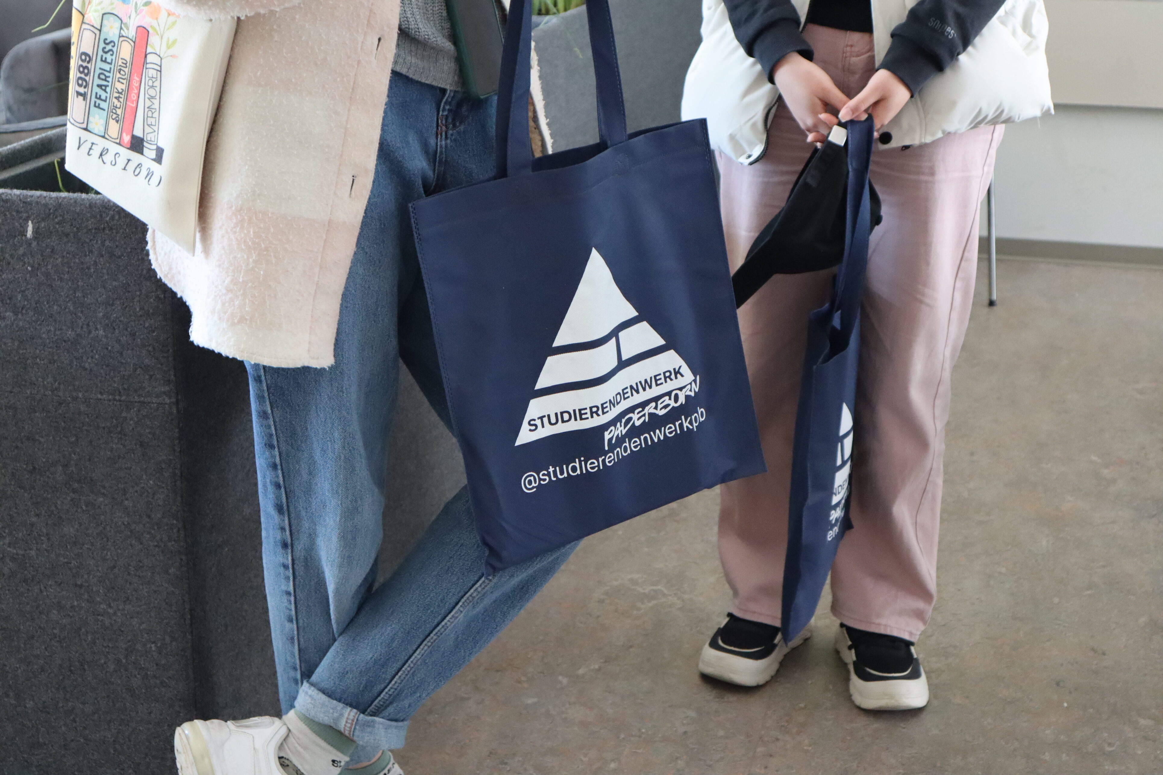 Two people are standing next to each other. They are each holding a blue bag with the triangular logo of the Paderborn Student Union.