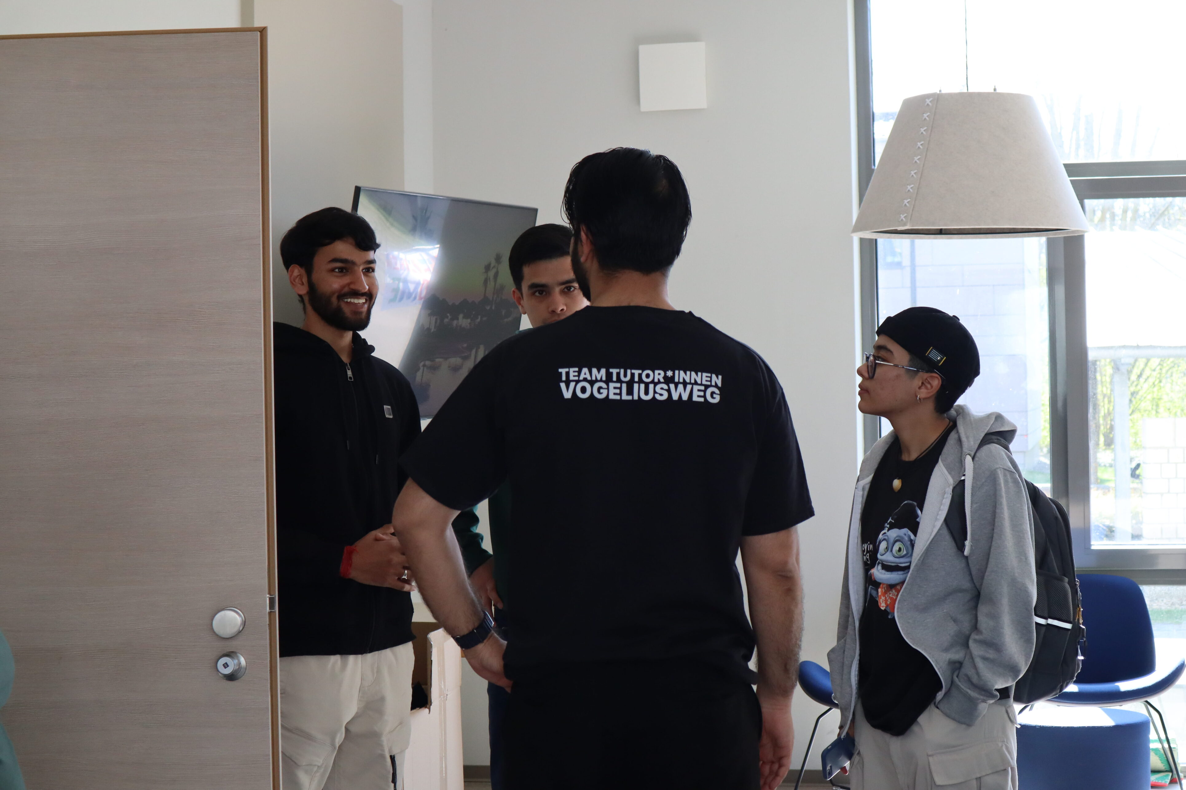 Vier Personen stehen im Kreis und unterhalten sich. Die Person, die mit dem Rücken zur Kamera gerichtet ist, trägt ein T-Shirt mit der Aufschrift Team Tutor*innen Vogeliusweg.