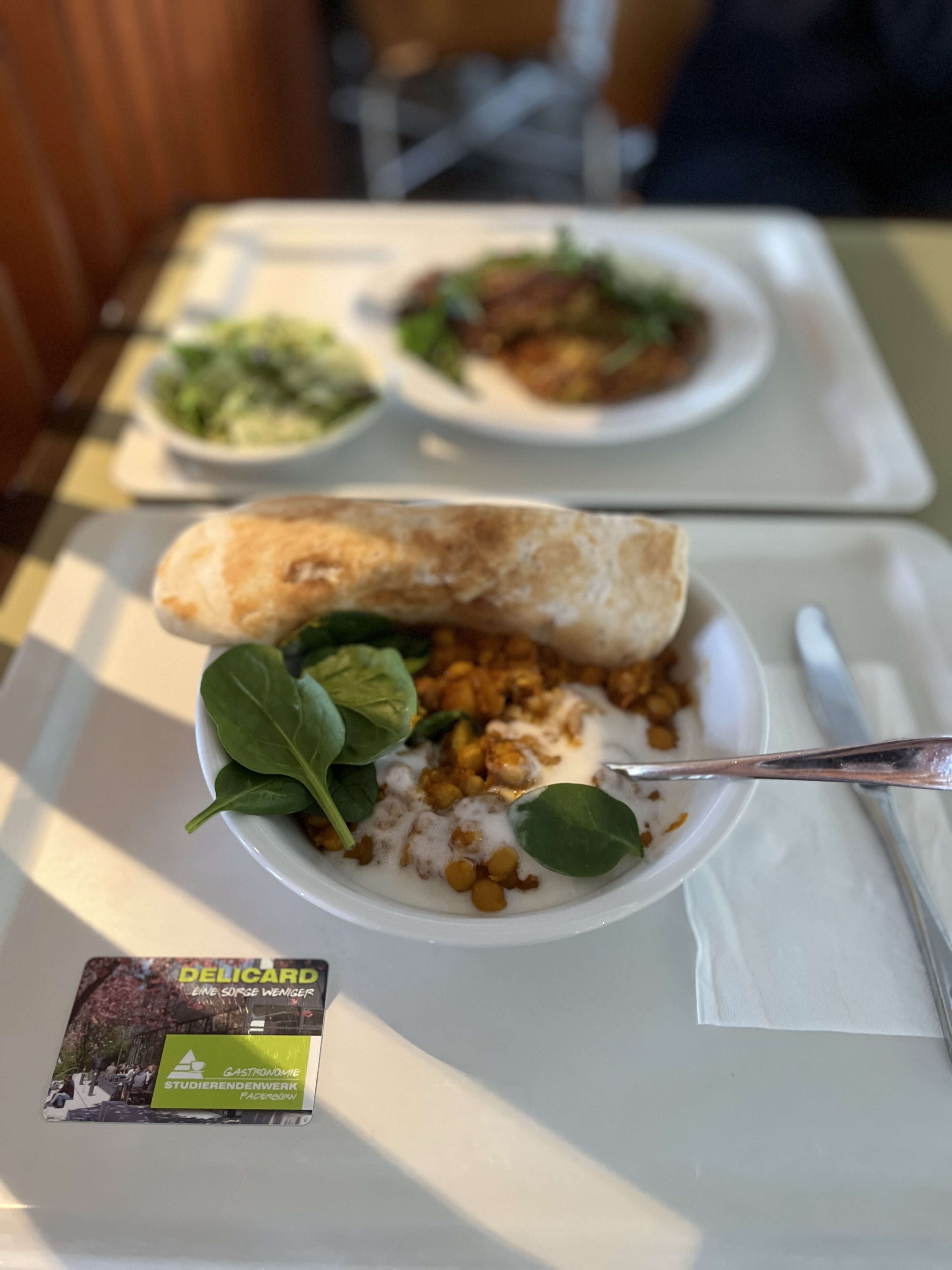 Zwei Tabletts liegen voreinander auf einem Tisch. Auf dem vorderen Tablett ist ein Teller mit einem Eintopf, Blattspinat und Naanbrot.