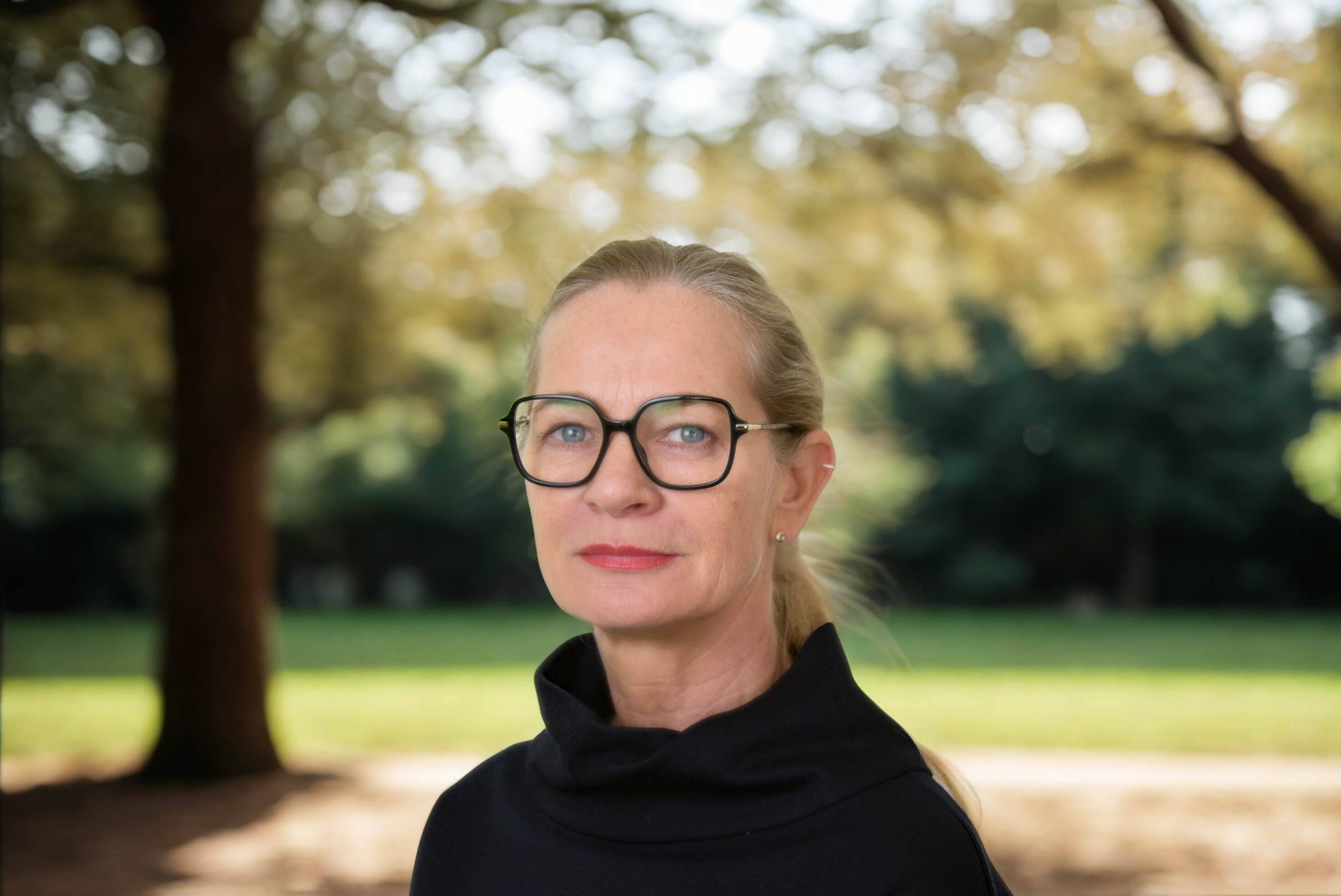 Eine Person steht draußen in der Natur und schaut in die Kamera. Sie trägt einen Zopf und eine Brille.