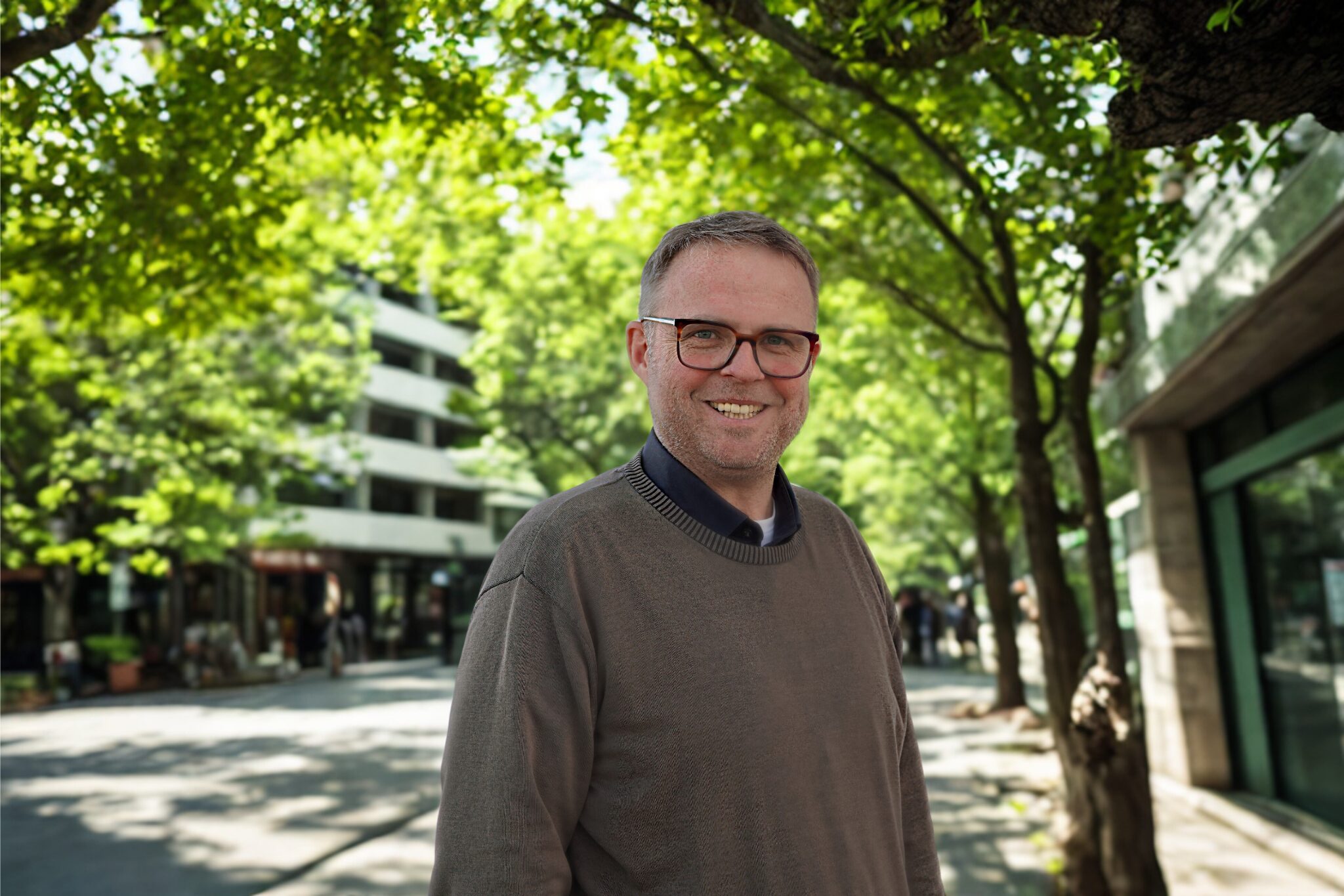 Eine Person steht vor einer Allee. Sie trägt ein dunkles Hemd mit einem braunen Pullover darüber.
