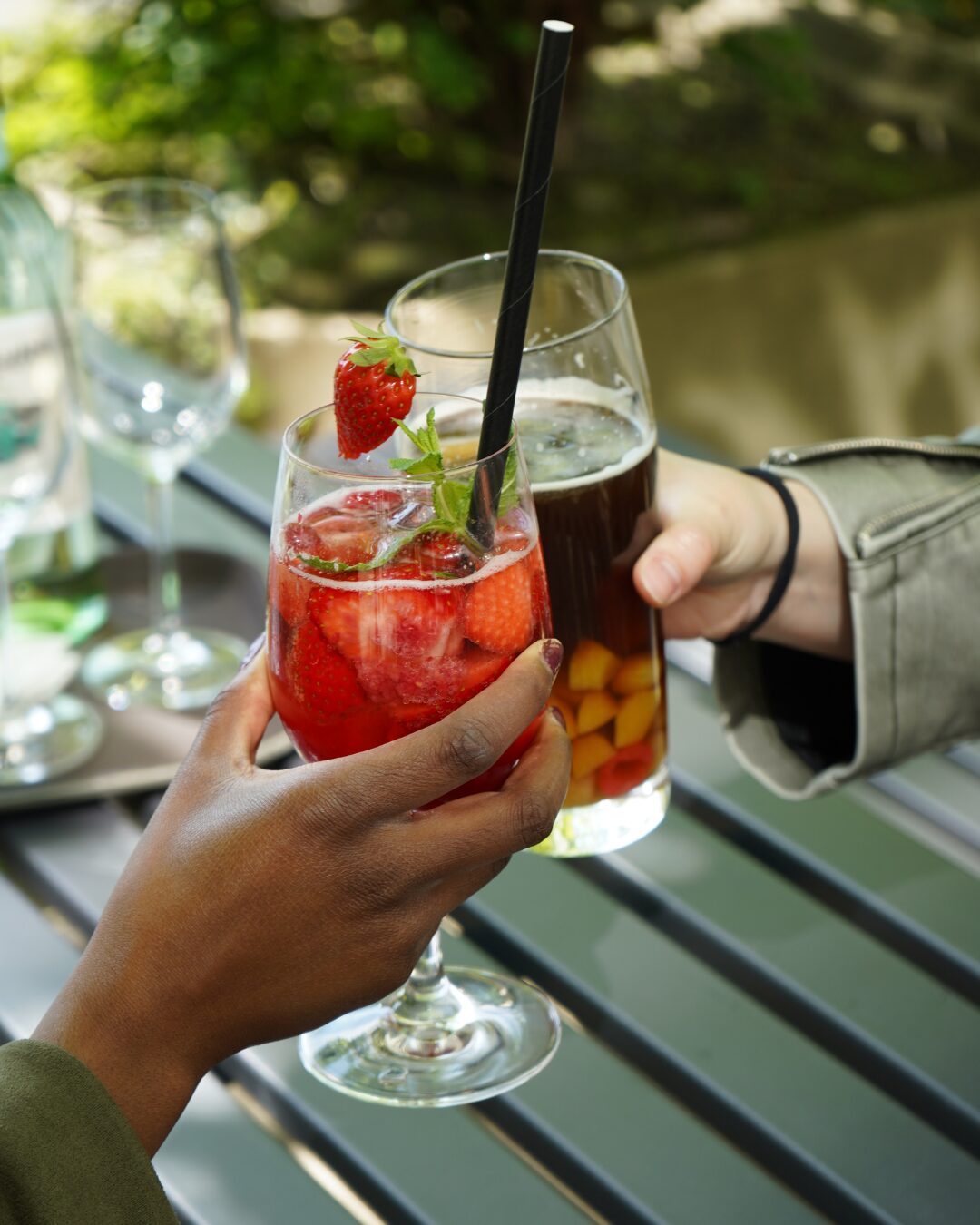 Zwei Hände halten jeweils ein Glas in der Hand, die aneinander gehalten werden. In dem einen Glas befindet sich ein rotes Getränk mit Erdbeeren und in dem anderen ein dunkles Getränk mit Früchten.