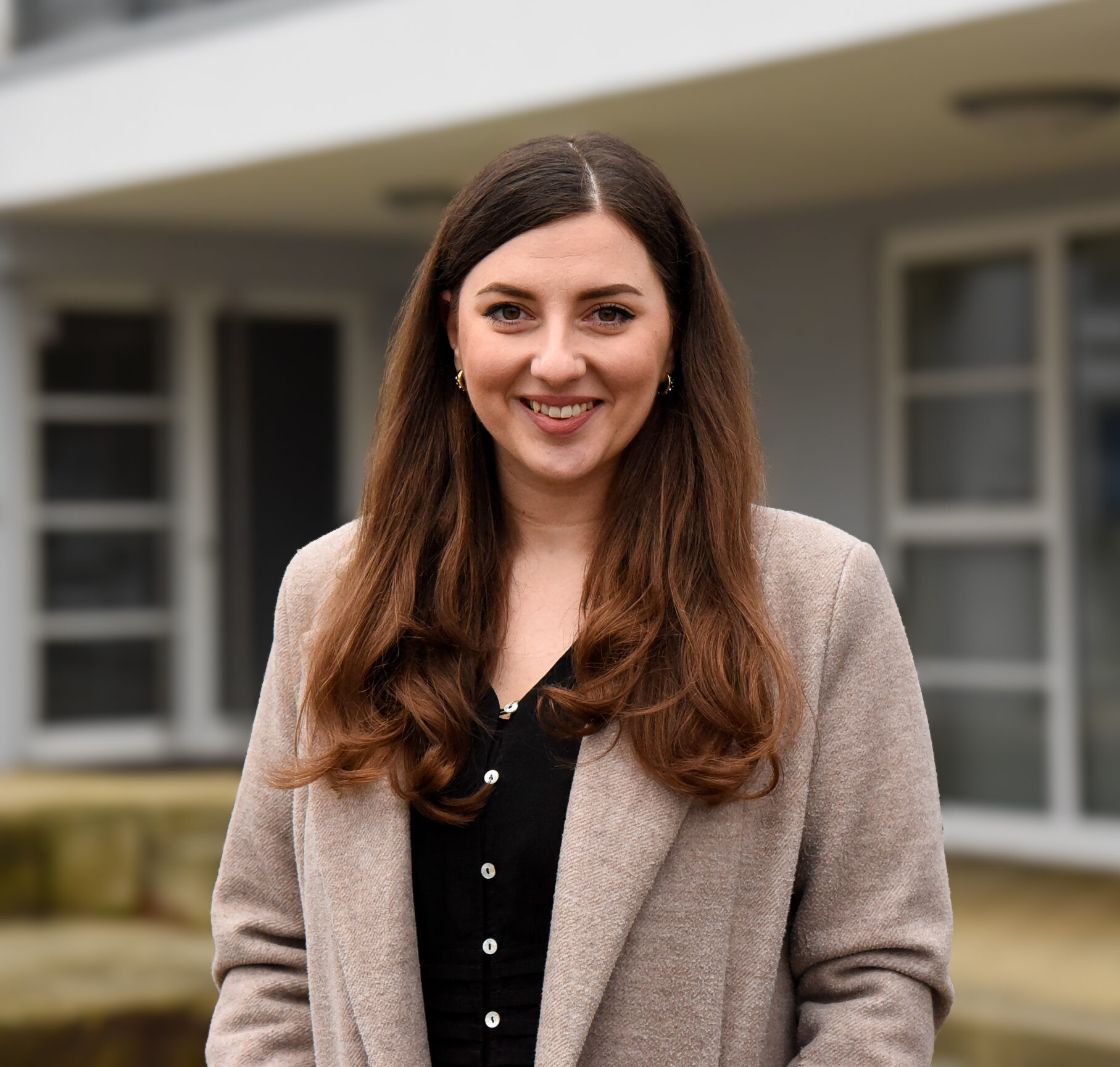 Eine Person mit langen braune Haaren und einem beigefarbenen Blazer steht vor dem Eingang eines Gebäudes.