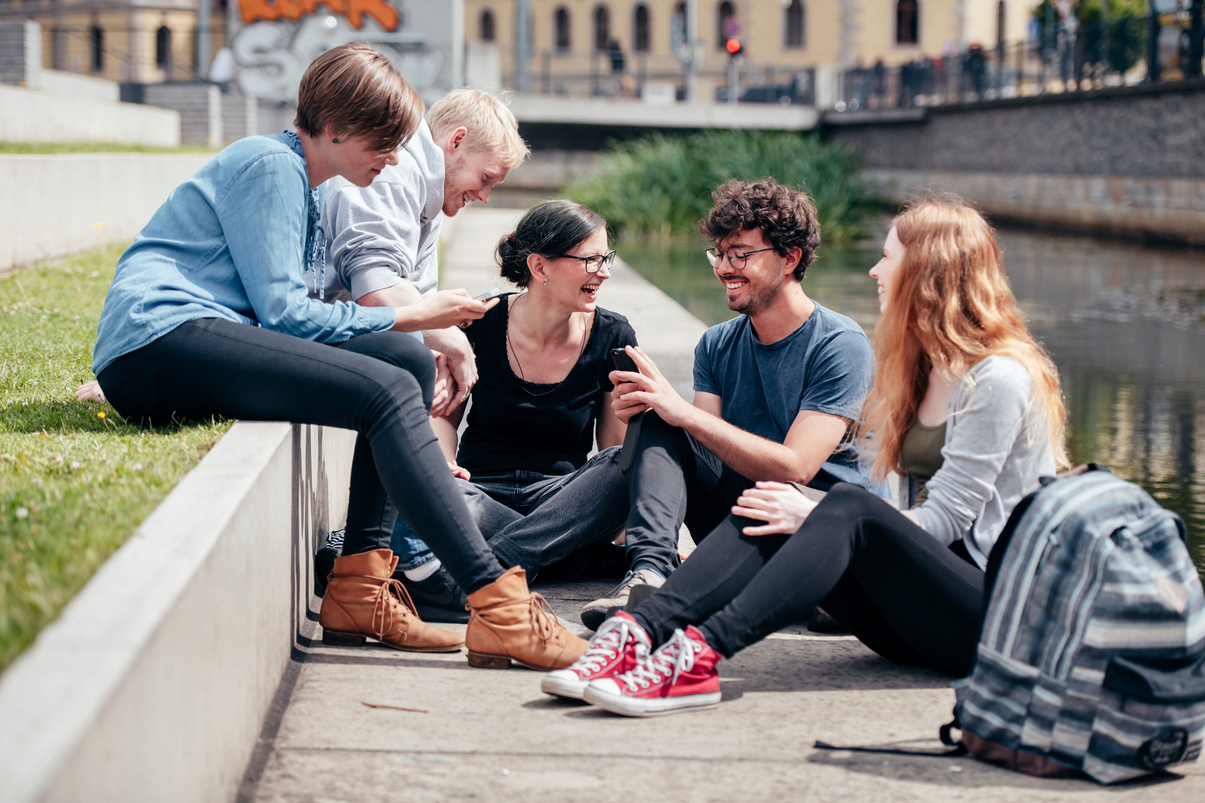 5 Personen, die bei gutem Wetter lachend zusammen auf einer kleinen Mauer und dem Gehweg davor sitzen.