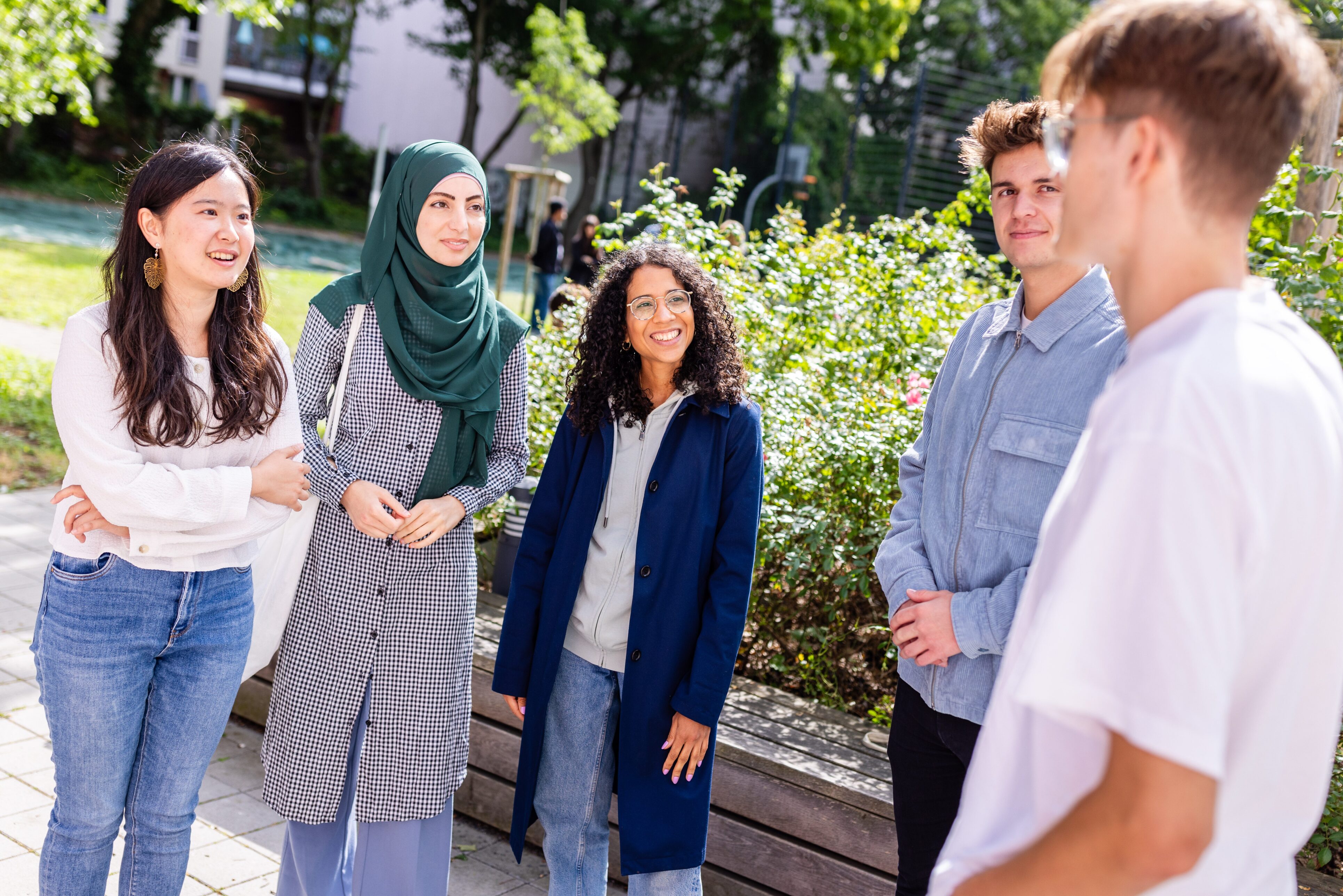 Fünf Personen stehen bei Sonnenschein draußen in einem Halbkreis und erzählen sich etwas.