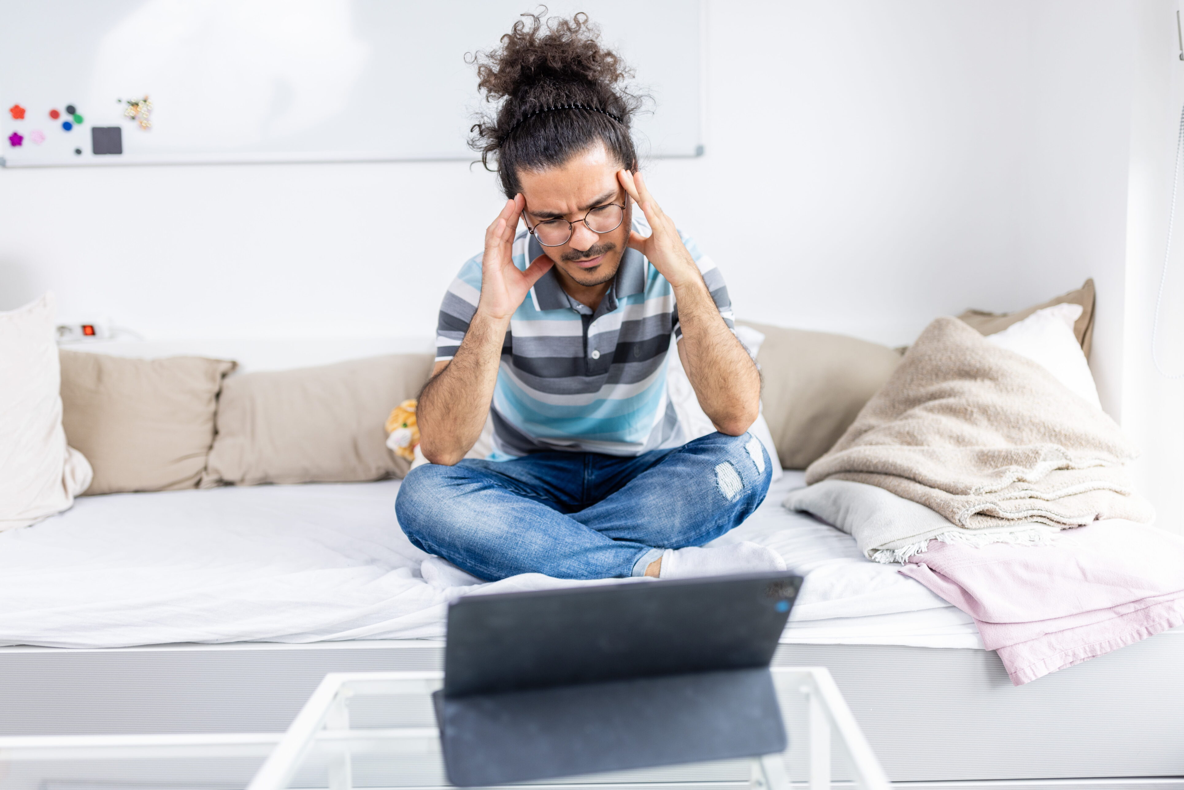Eine Person sitzt auf einem Sofa und schaut auf ein Tablet. Dabei fasst sie sich mit beiden Händen an den Kopf.