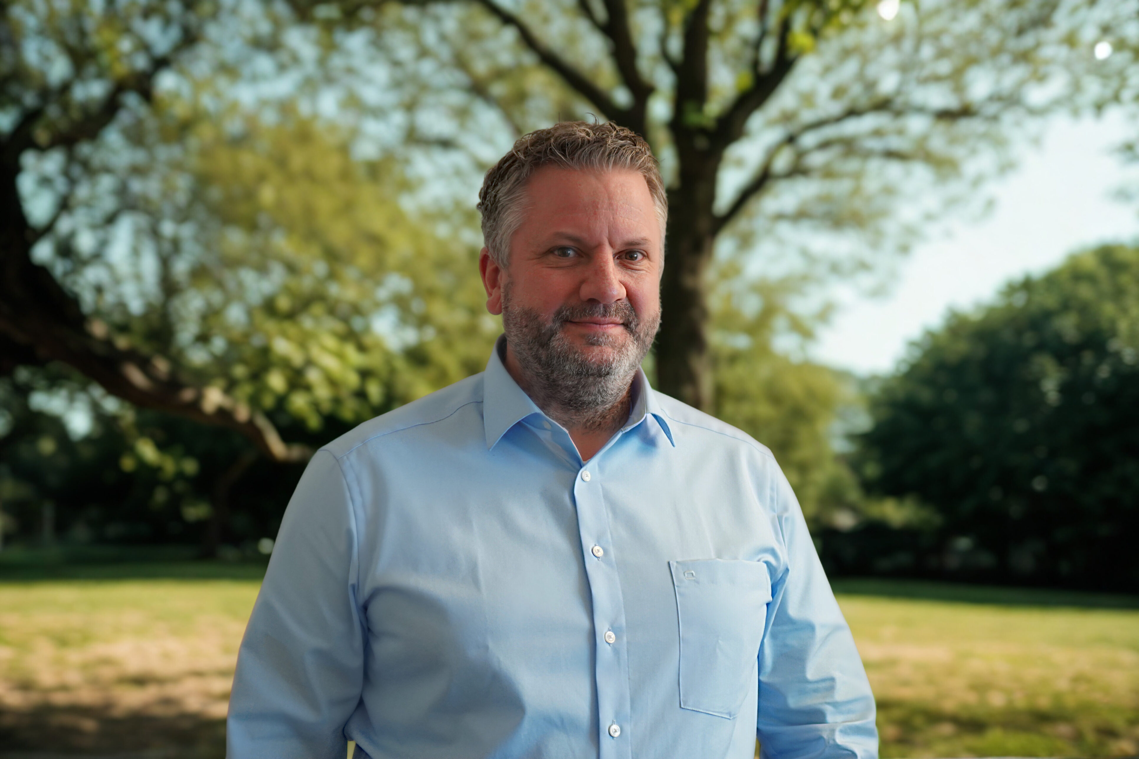 Eine Person steht vor einem Baum und schaut in die Kamera. Sie trägt einen Bart und ein Hemd.