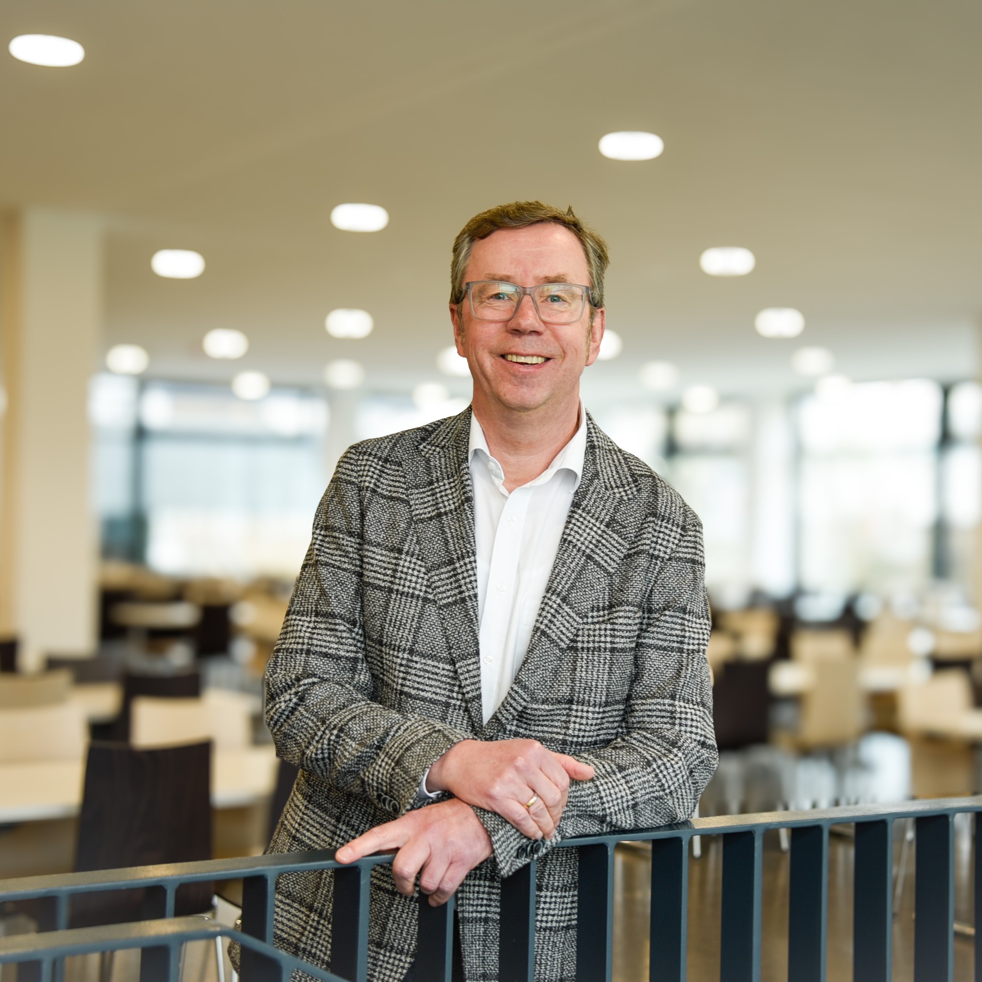 Eine Person mit Brille und Sakko lächelt in die Kamera und lehnt dabei an einem Geländer.