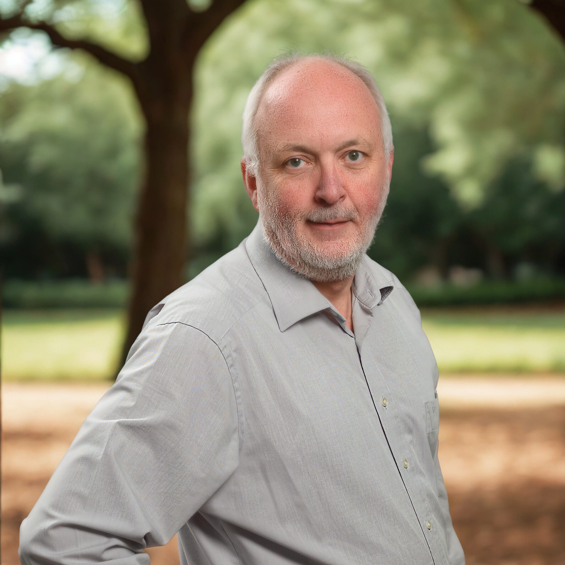 Eine Person steht im Schatten unter einem Baum und schaut in die Kamera. Sie trägt ein graues Hemd und hat den rechten Arm in die Hüfte gestützt.