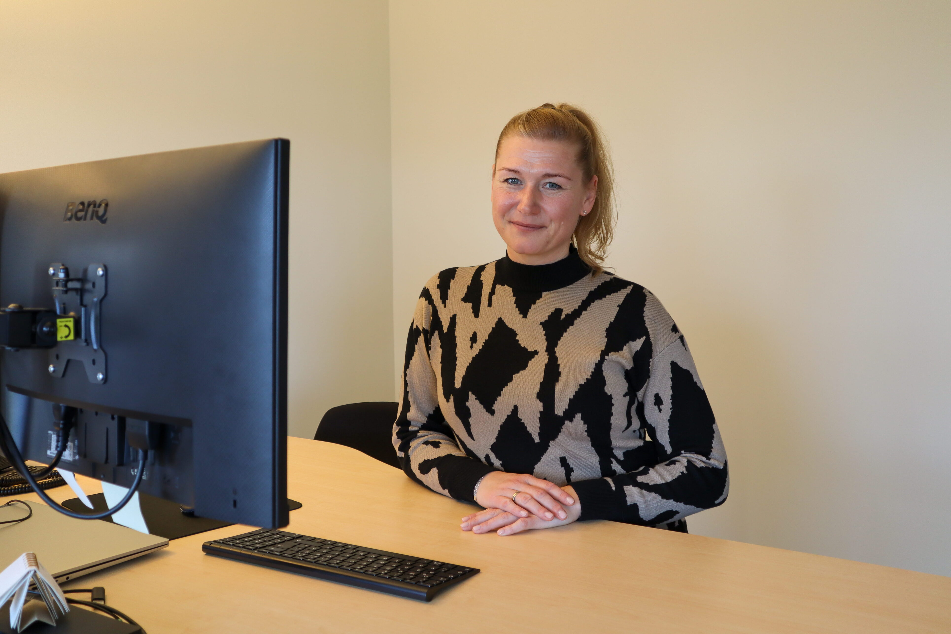 Eine Person sitzt vor einem Bildschirm und hat ihre Hände vor sich auf dem Schreibtisch abgelegt. Sie trägt einen Zopf und einen Schwarz-Beige gemusterten Pullover.