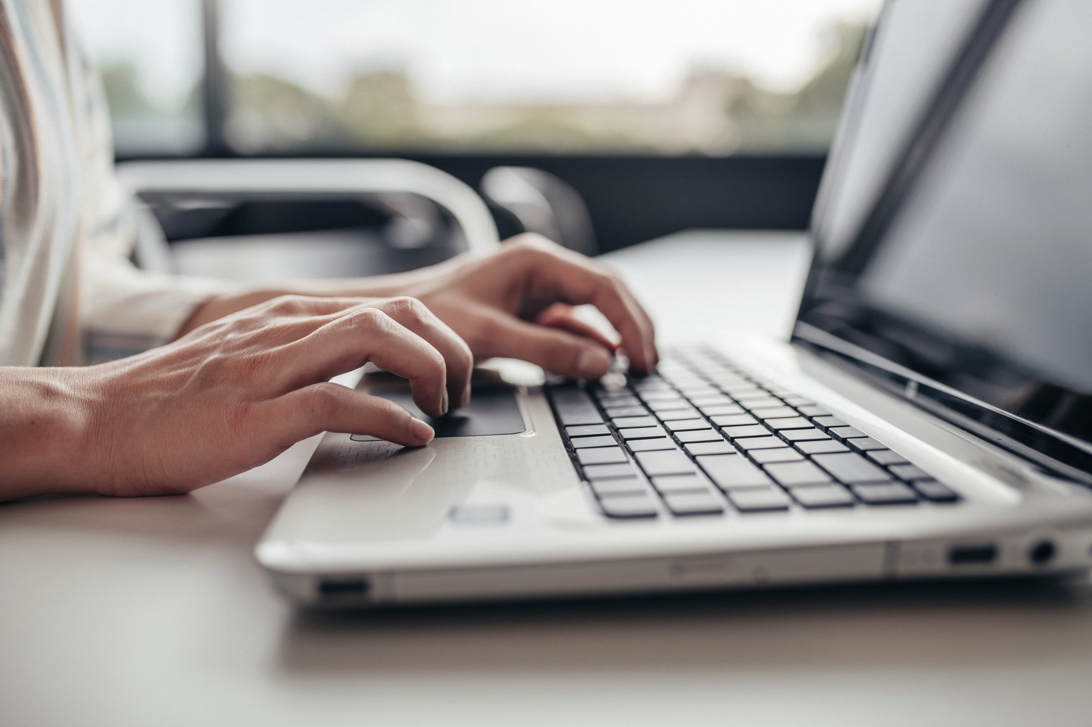 Hände tippen auf der Tastatur eines Laptops, der auf einem Tisch steht, mit einem großen Fenster im Hintergrund.