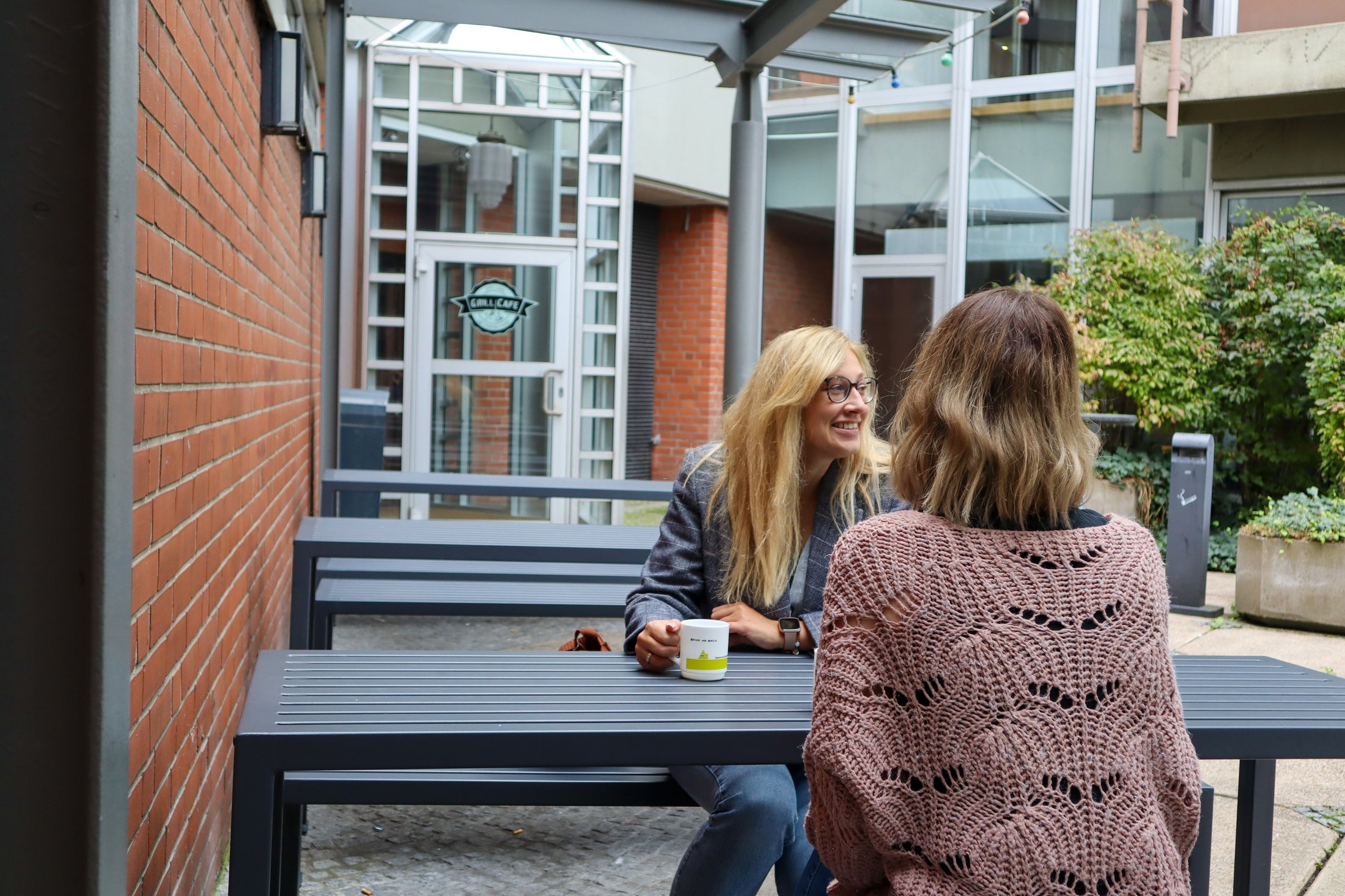 Zwei Personen sitzen an einem Tisch im Freien, umgeben von einem begrünten Innenhof und modernen Gebäuden.