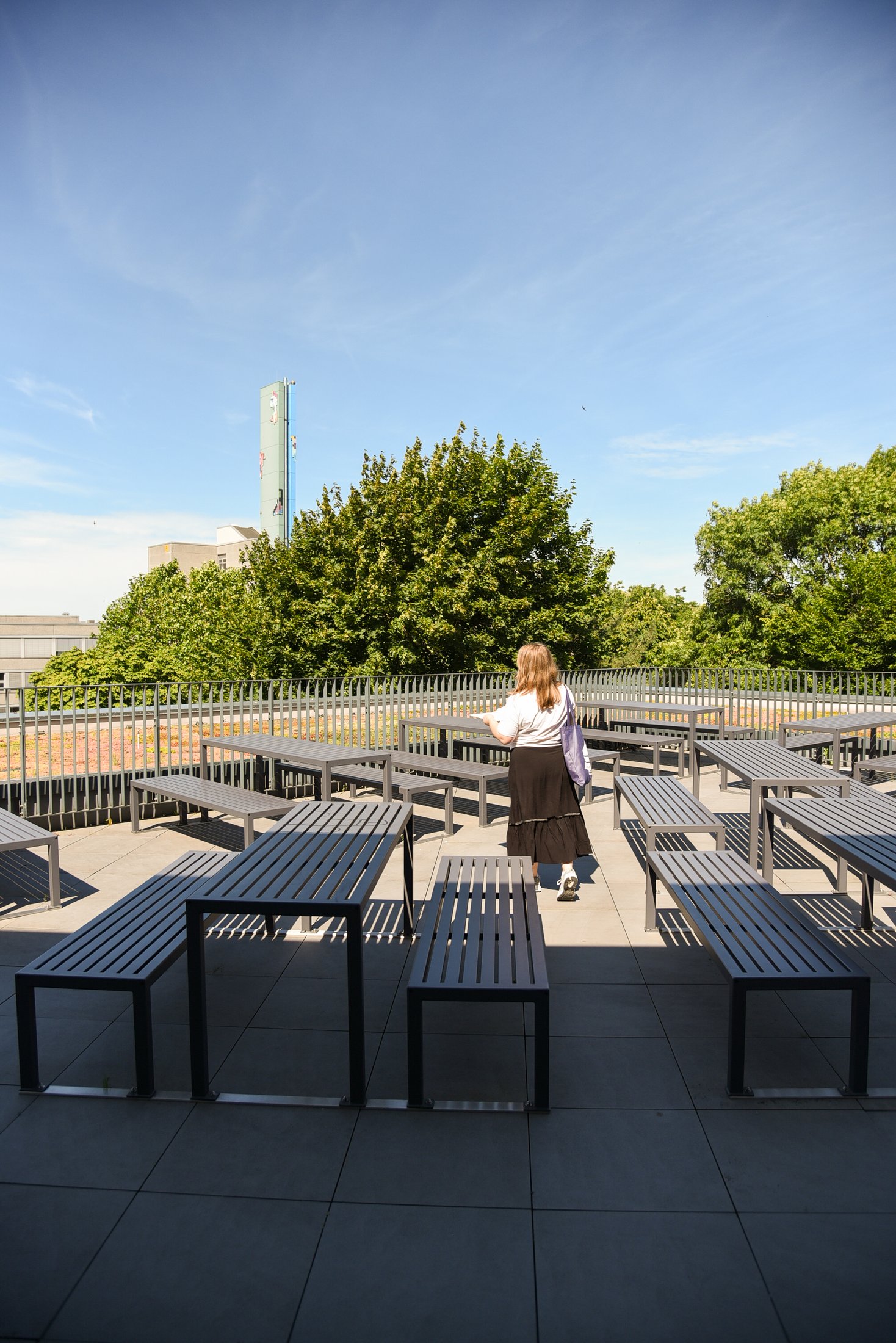 Eine sonnige Terrasse mit grauen Tischen und Bänken. Im Hintergrund ist eine Grünfläche und Bäume zu sehen.