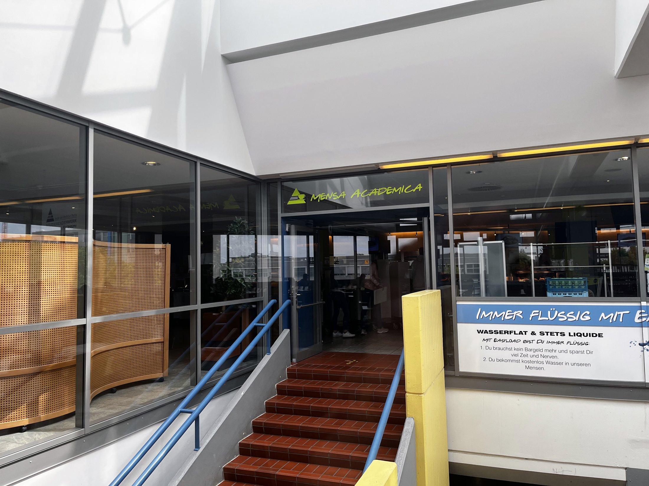 Eingangsbereich einer Mensa mit großem Glasfenster, Holztisch und Hinweisschildern zum Wasserangebot.