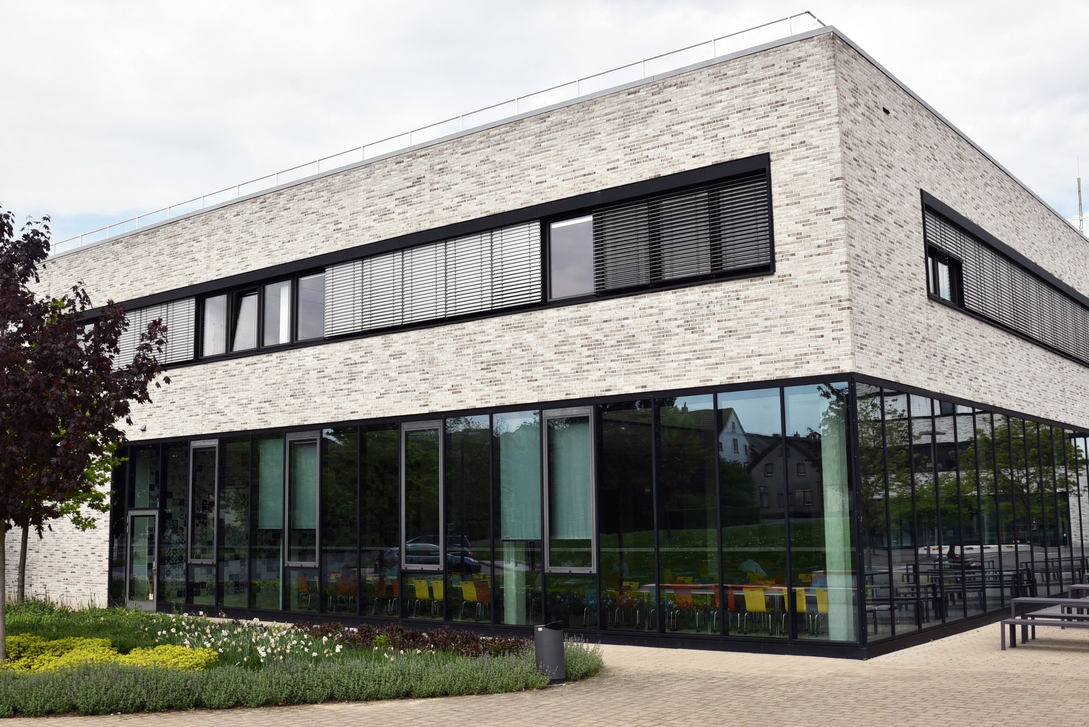 Moderne Mensa in Lippstadt mit großflächigen Fensterfronten, grüner Bepflanzung und bunten Stühlen im Außenbereich.