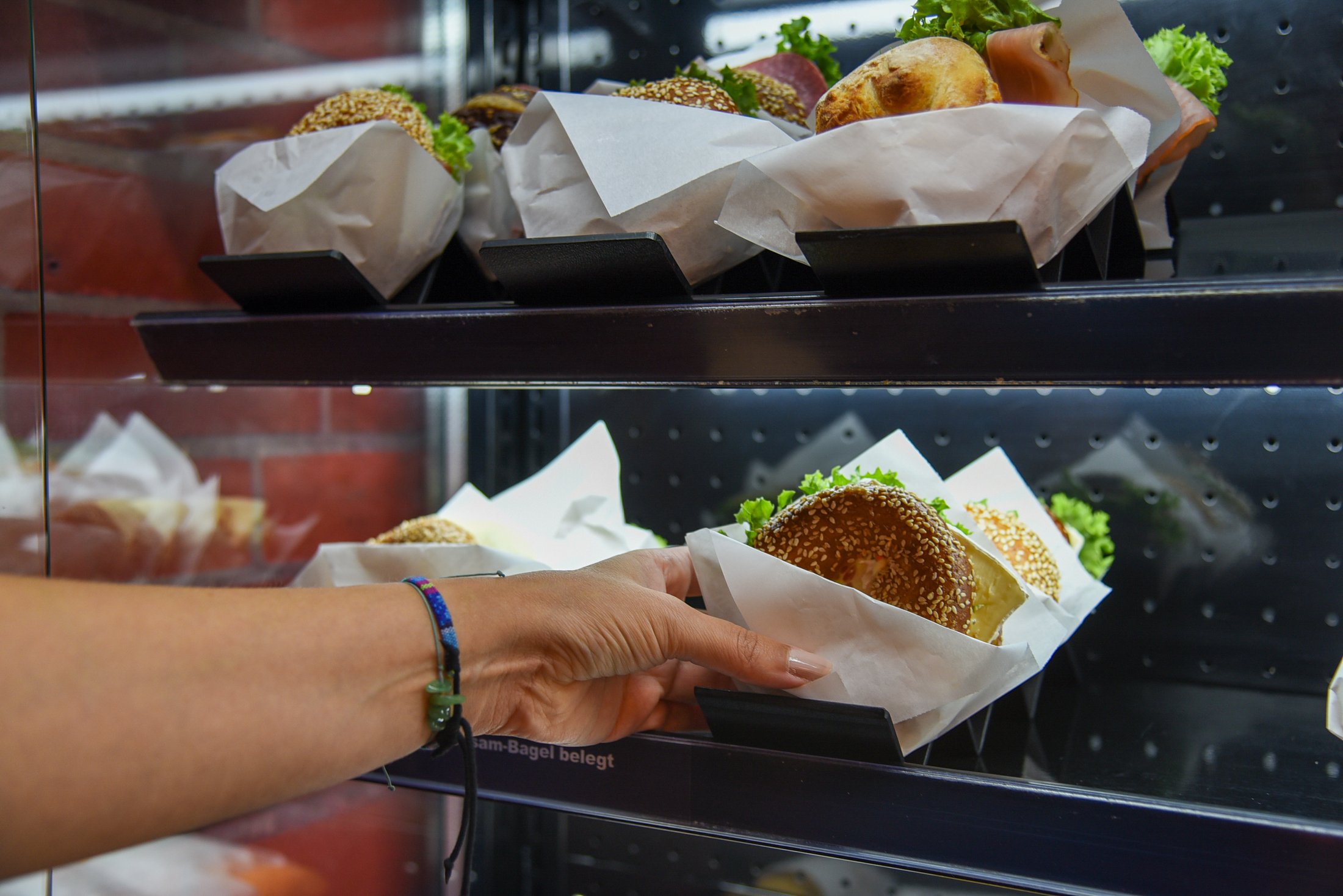 Hand greift nach einem belegten Bagel in einem Verkaufsregal mit mehreren Bagels, die in Papier eingeschlagen sind.