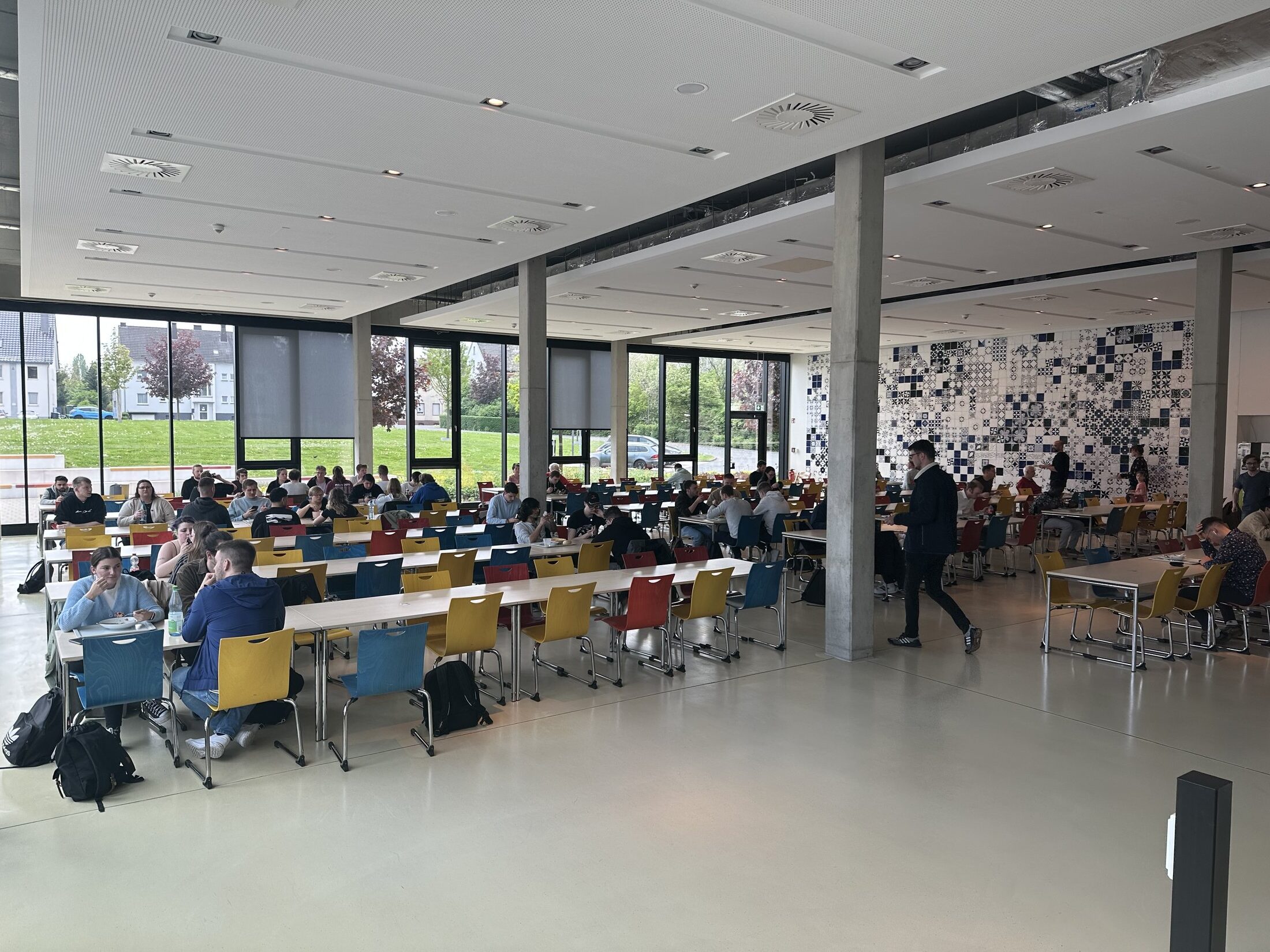 Bunte Stühle und Tische in einer Mensa; Menschen sitzen und essen, Fenster mit Blick ins Freie. Grafische Wanddekoration.