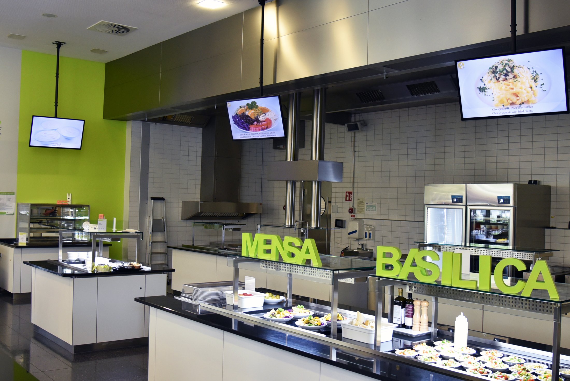 Moderne Mensa mit Essensausgabe, großen Theken, frischen Salaten und digitalen Displays. Grüne Elemente im Design.