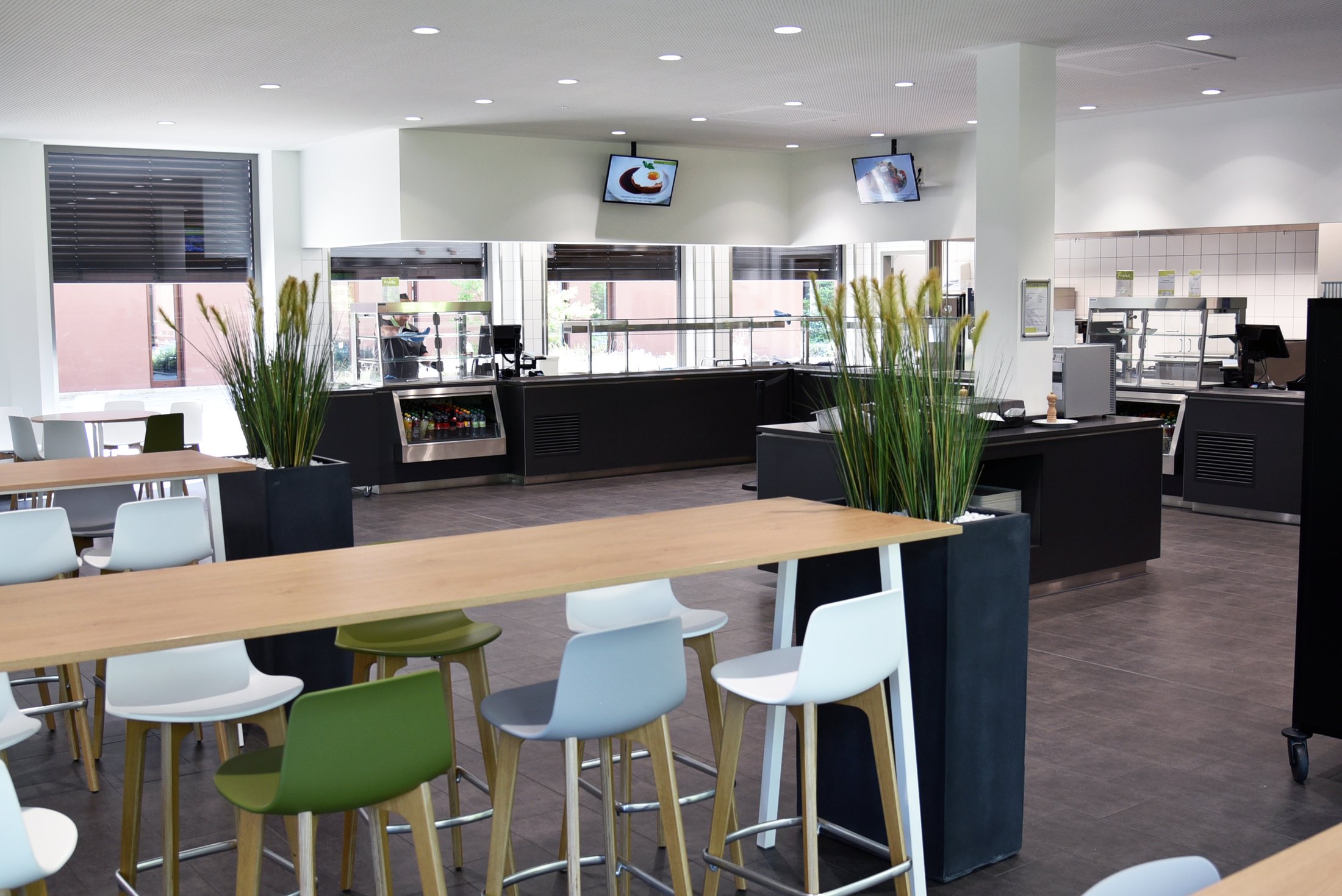 Eine moderne Mensa mit Sitzbereichen, Stehtischen, Pflanzen und einer offenen Küche im Hintergrund.