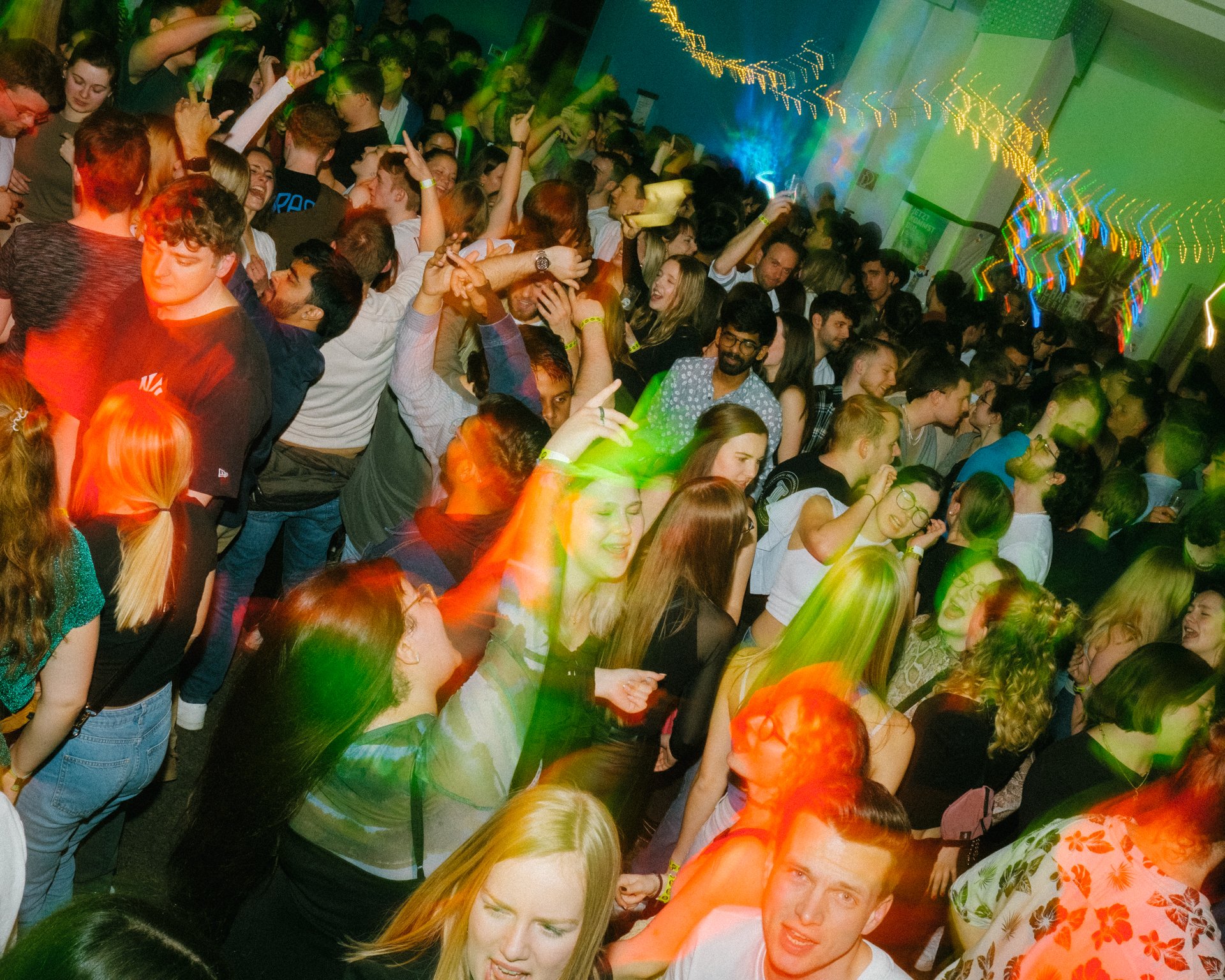 Menschenmenge tanzt in einem Raum mit grellen Lichtern und festlicher Stimmung, Musik und Bewegung prägen die Szene.