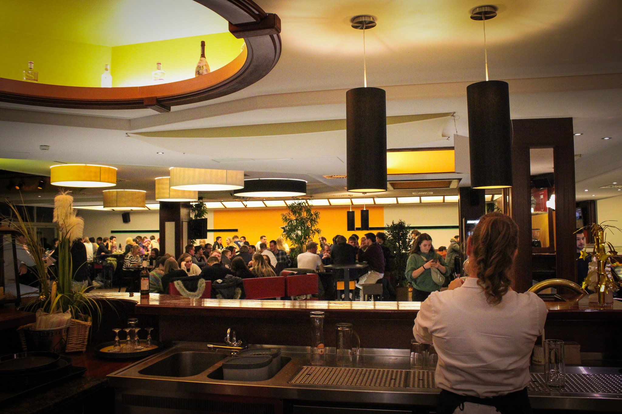 Innenansicht einer Gastronomie mit modernen Lampen, Käufern an Tischen und einem Barbereich im Vordergrund.