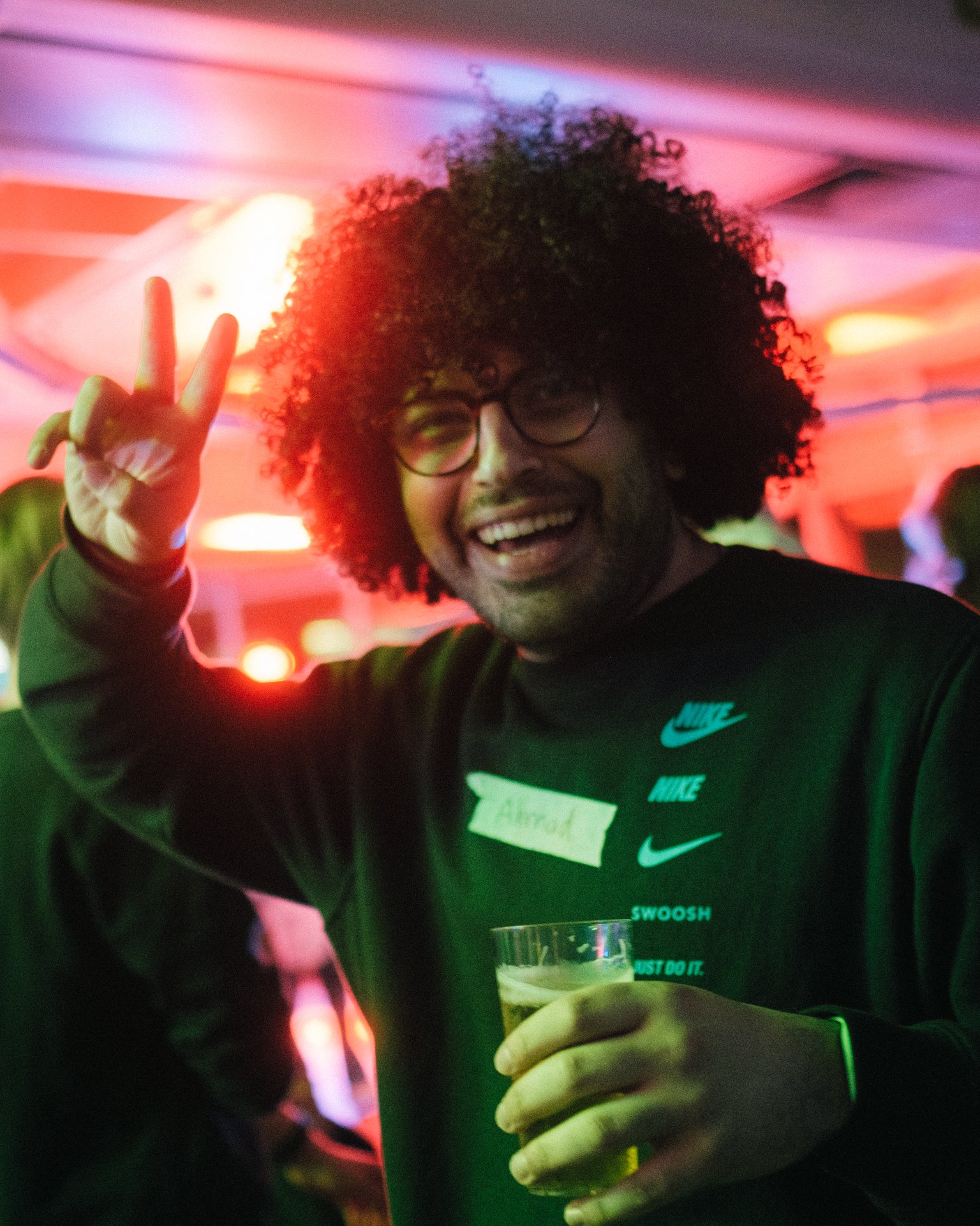 Ein fröhlicher Mensch mit lockigem Haar zeigt das Peace-Zeichen und hält ein Getränk, umgeben von bunten Lichtern.