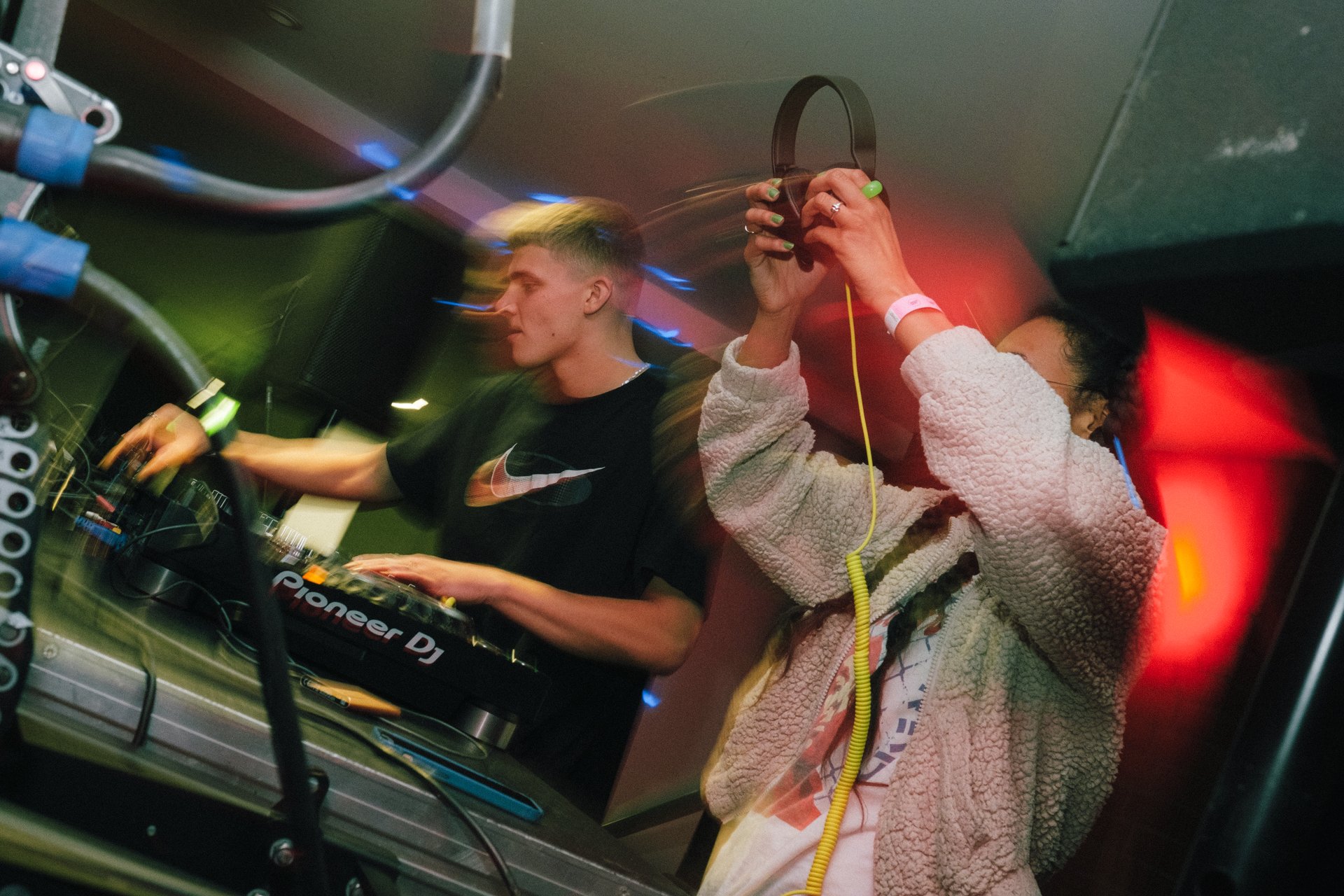 Zwei DJs stehen an einem Mischpult, umgeben von bunten Lichtern und Kabeln, die Musik auflegen.