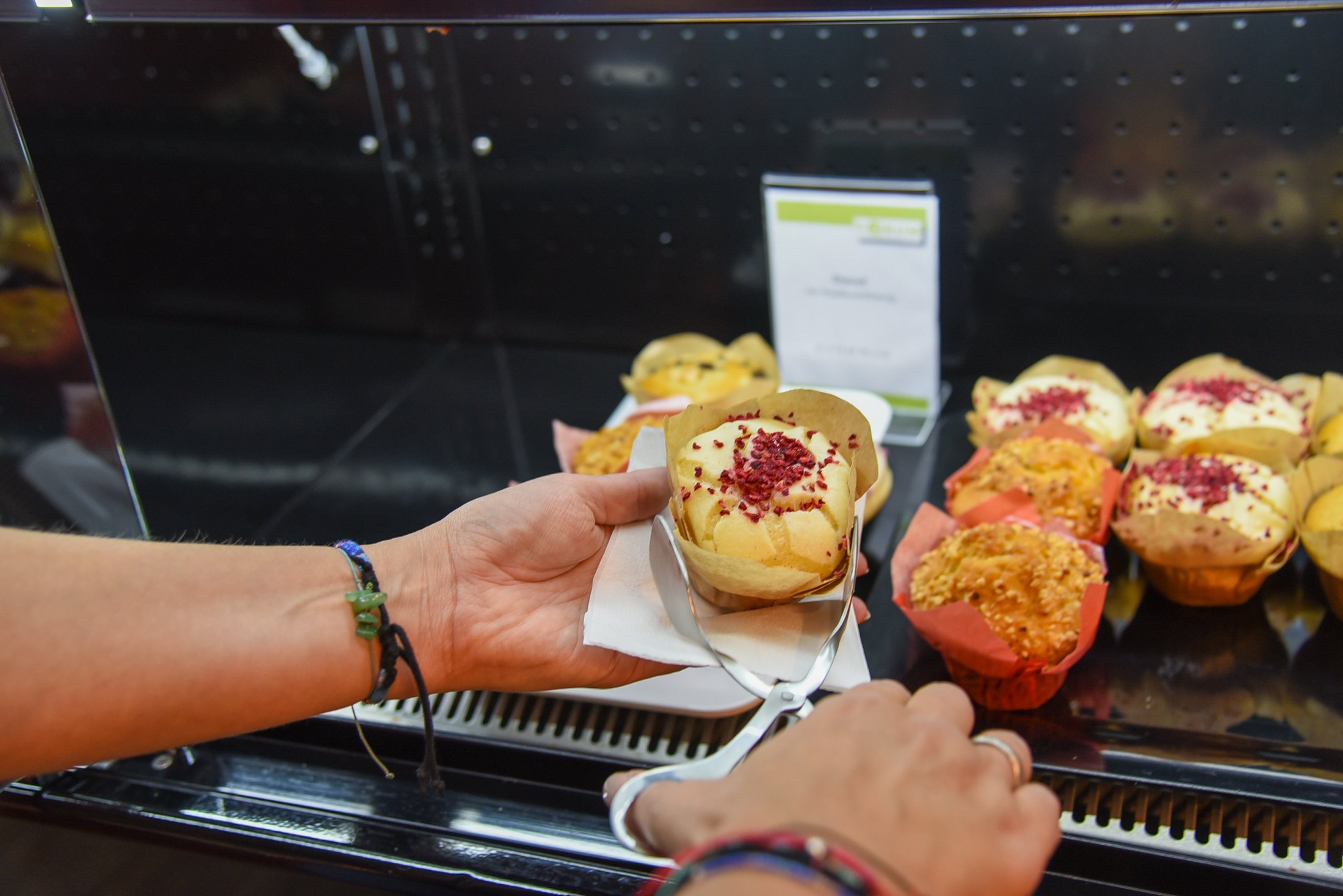 Ein Muffin mit Frischkäse und Dekor auf einem Teller wird mit einer Zange aus einer Theke genommen.