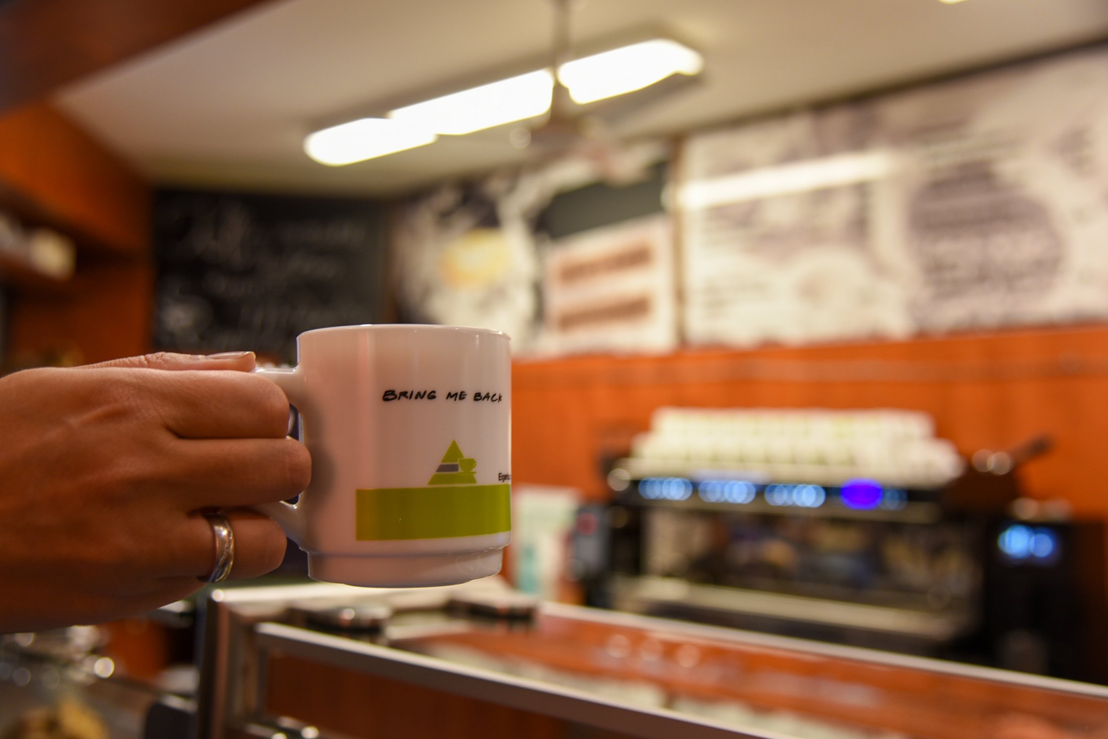 Tasse mit der Aufschrift "BRING ME BACK" und stilisiertem Logo, im Hintergrund eine Kaffeemaschine und Bar.