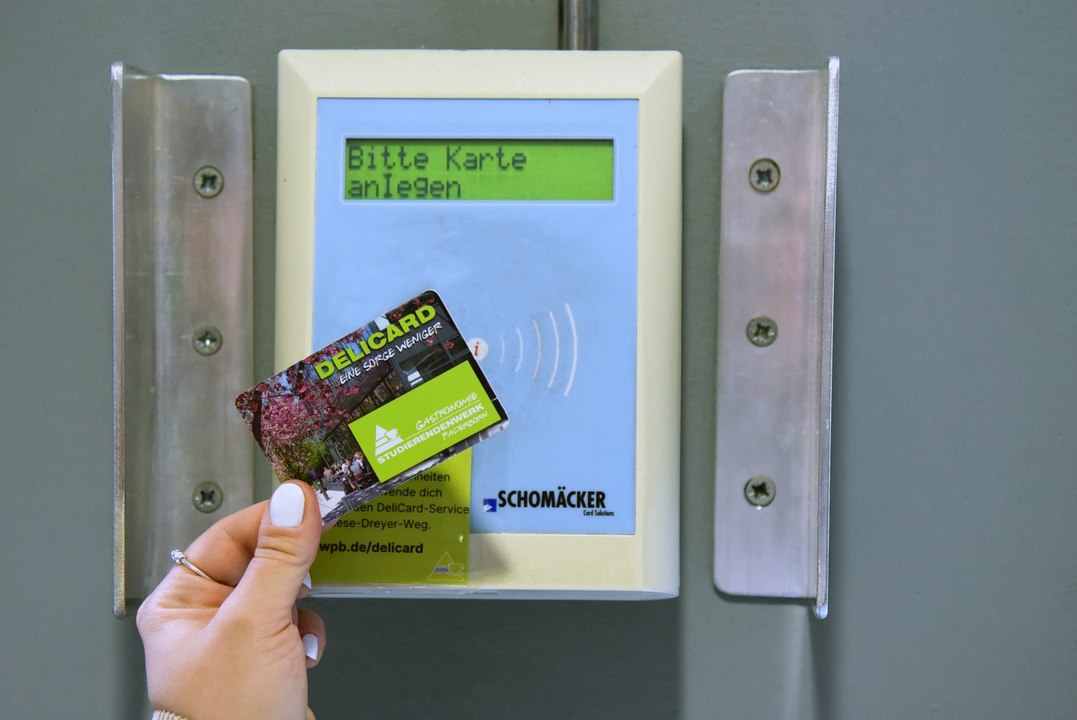 Eine Hand hält die DeliCard an ein Kartenlesegerät, auf dem die Aufforderung „Bitte Karte anlegen“ angezeigt wird.