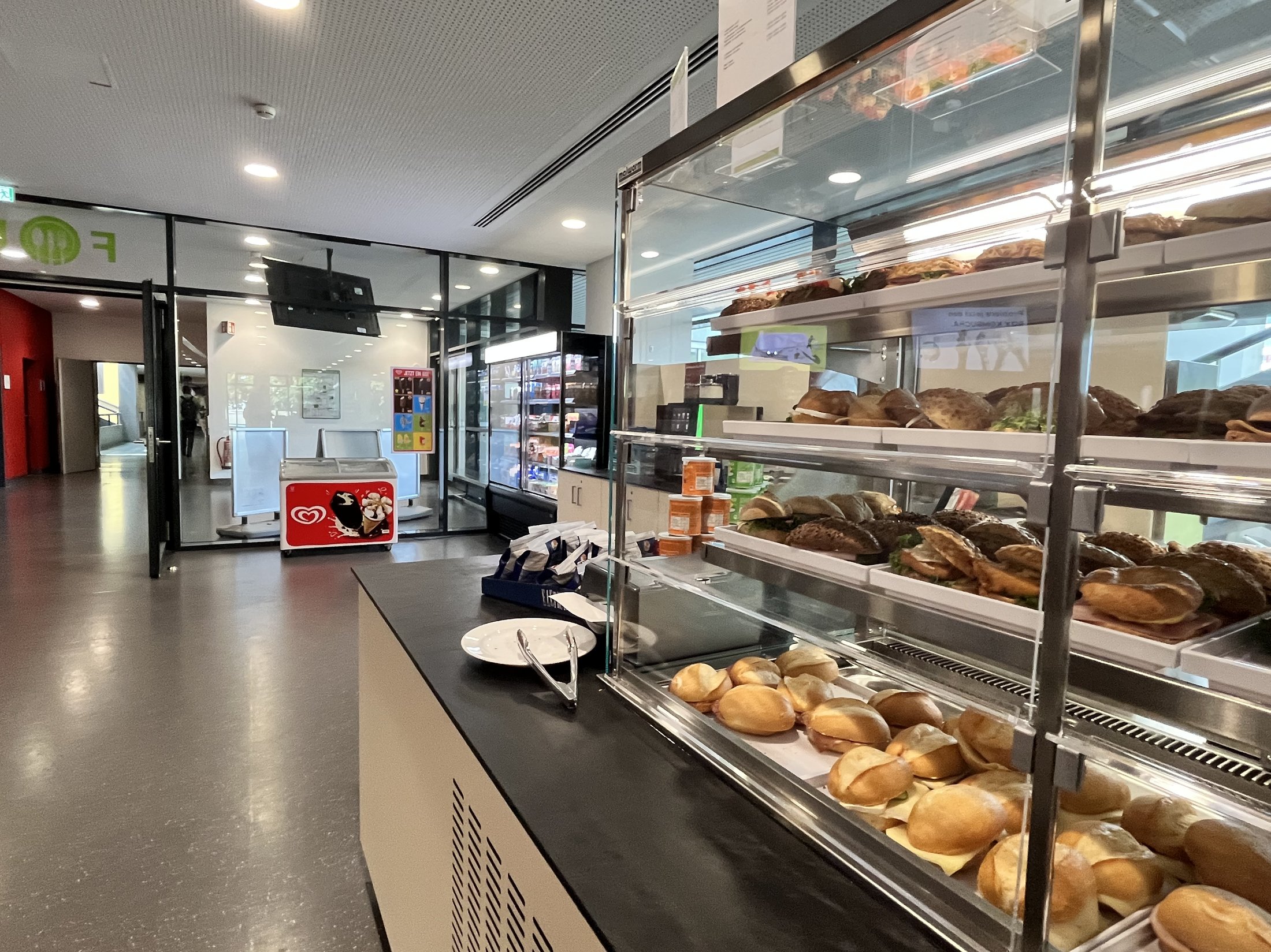 Ein einfacher Verkaufsbereich mit verschiedenen Brötchen und Snacks, Glasvitrine, im Hintergrund Automaten und eine große Fensterfront.