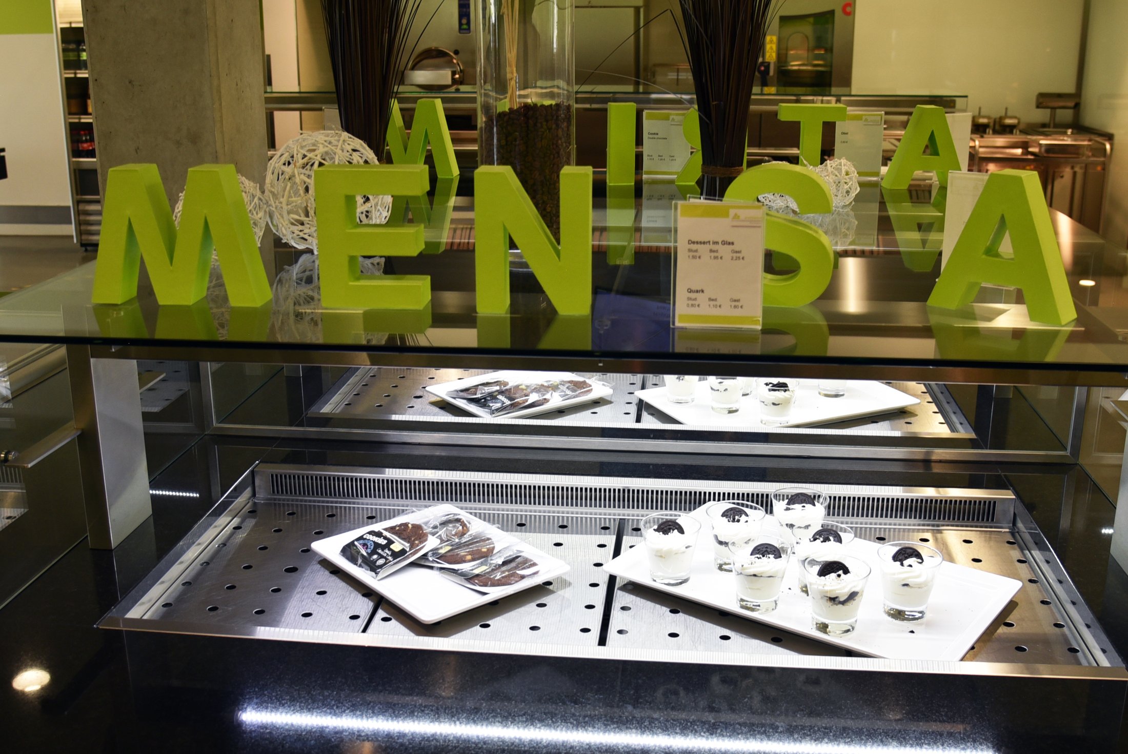 Grüne Buchstaben formen das Wort "MENSA". Auf einem Regal sind Desserts in Gläsern und Platten mit Keksen sichtbar.