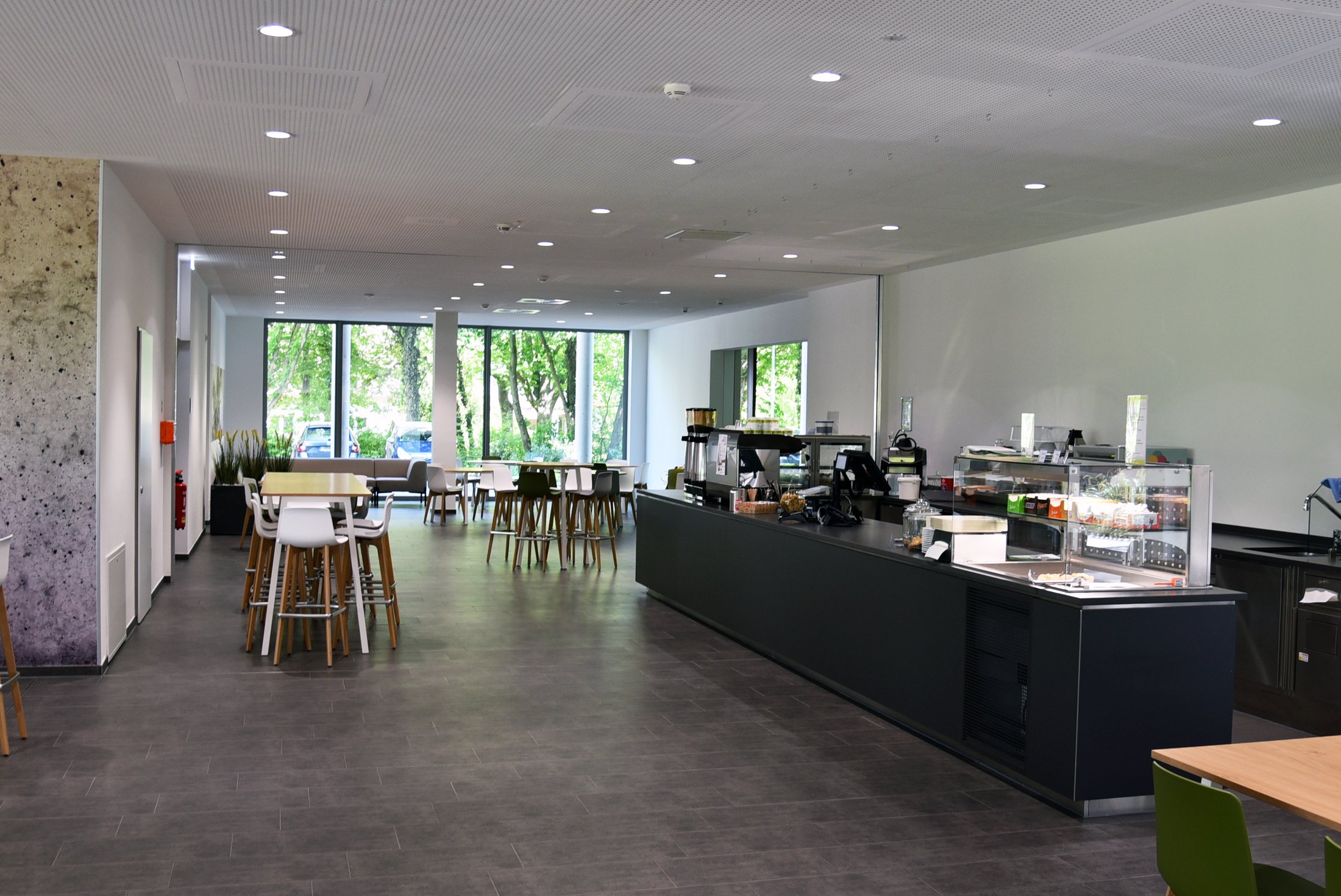 Großer, heller Gastronomiebereich mit Tischen, Stühlen und einem langen Tresen, der Speisen und Getränke anbietet.