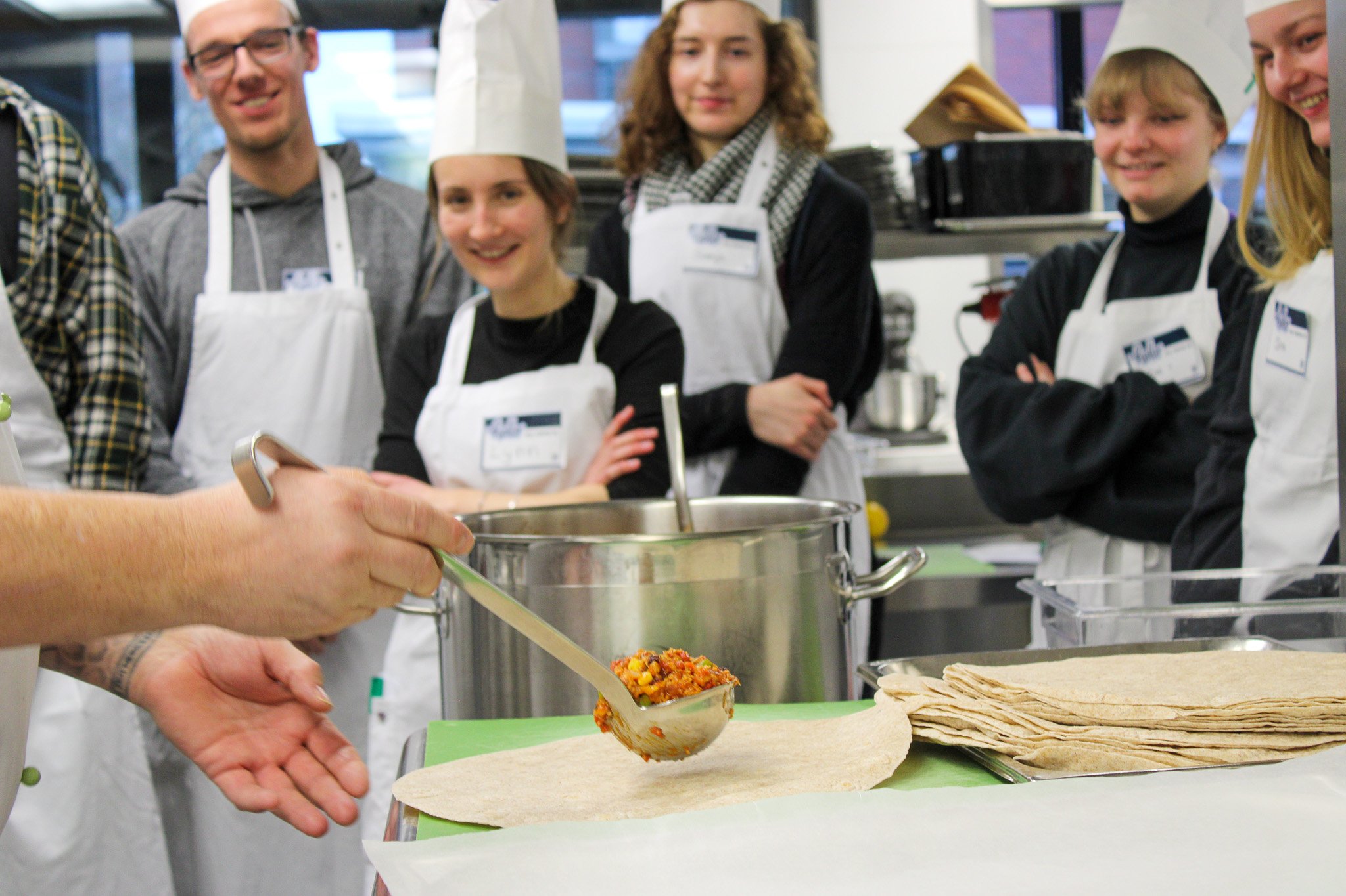 A group of people in chef's aprons and hats watch attentively while one person places filling on a flatbread with a spoon.