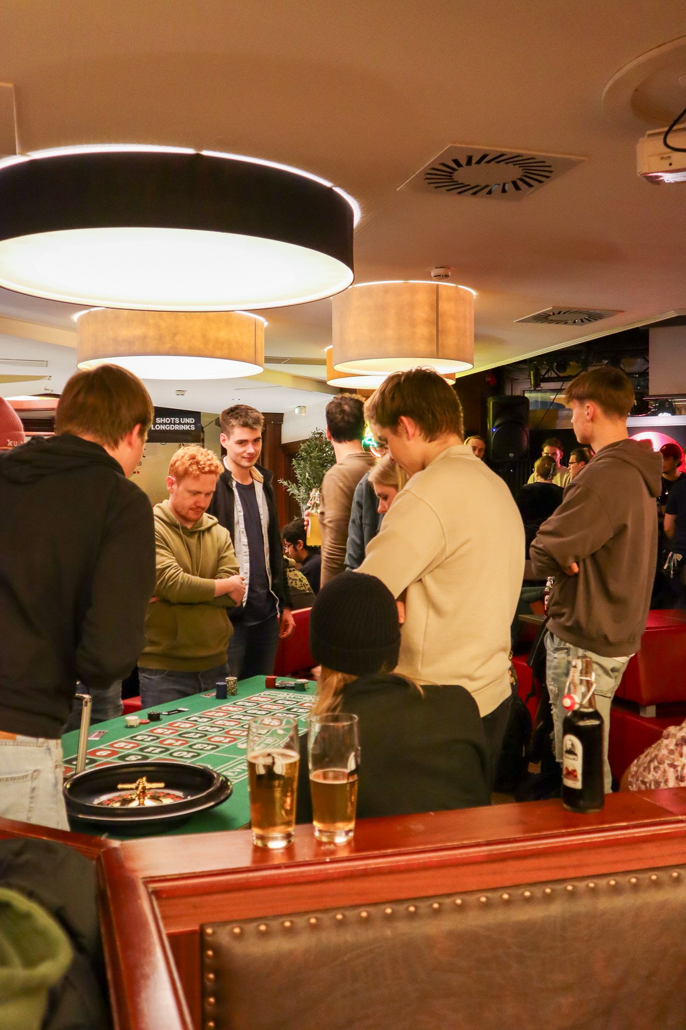 Gruppe von Menschen um einen Tisch mit einem Roulette-Spiel, in einer gut beleuchteten Bar mit Getränken.