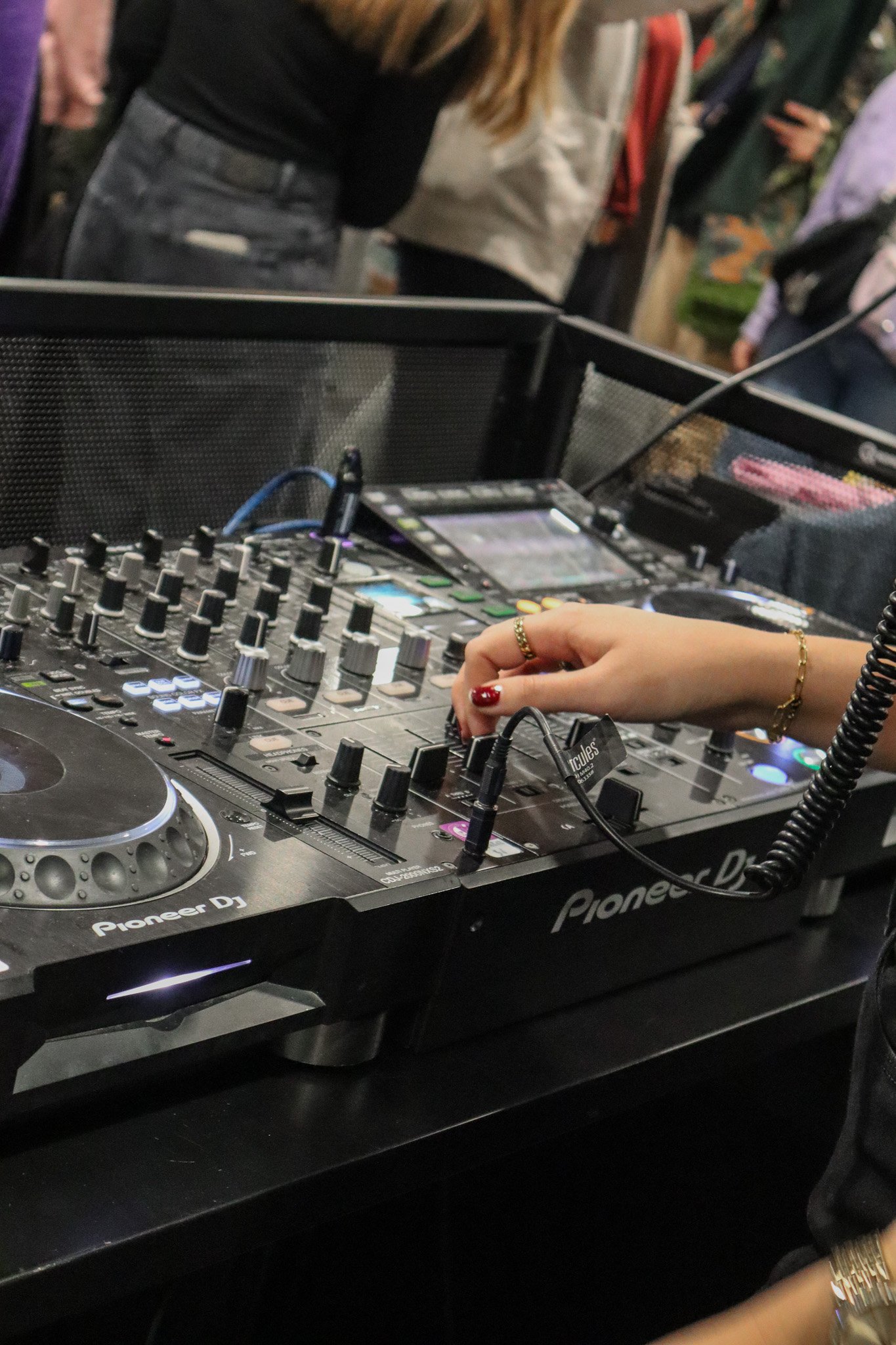 Mixer und DJ-Equipment sind im Vordergrund sichtbar. Eine Hand justiert einen Regler. Menschen im Hintergrund sind schemenhaft zu erkennen.